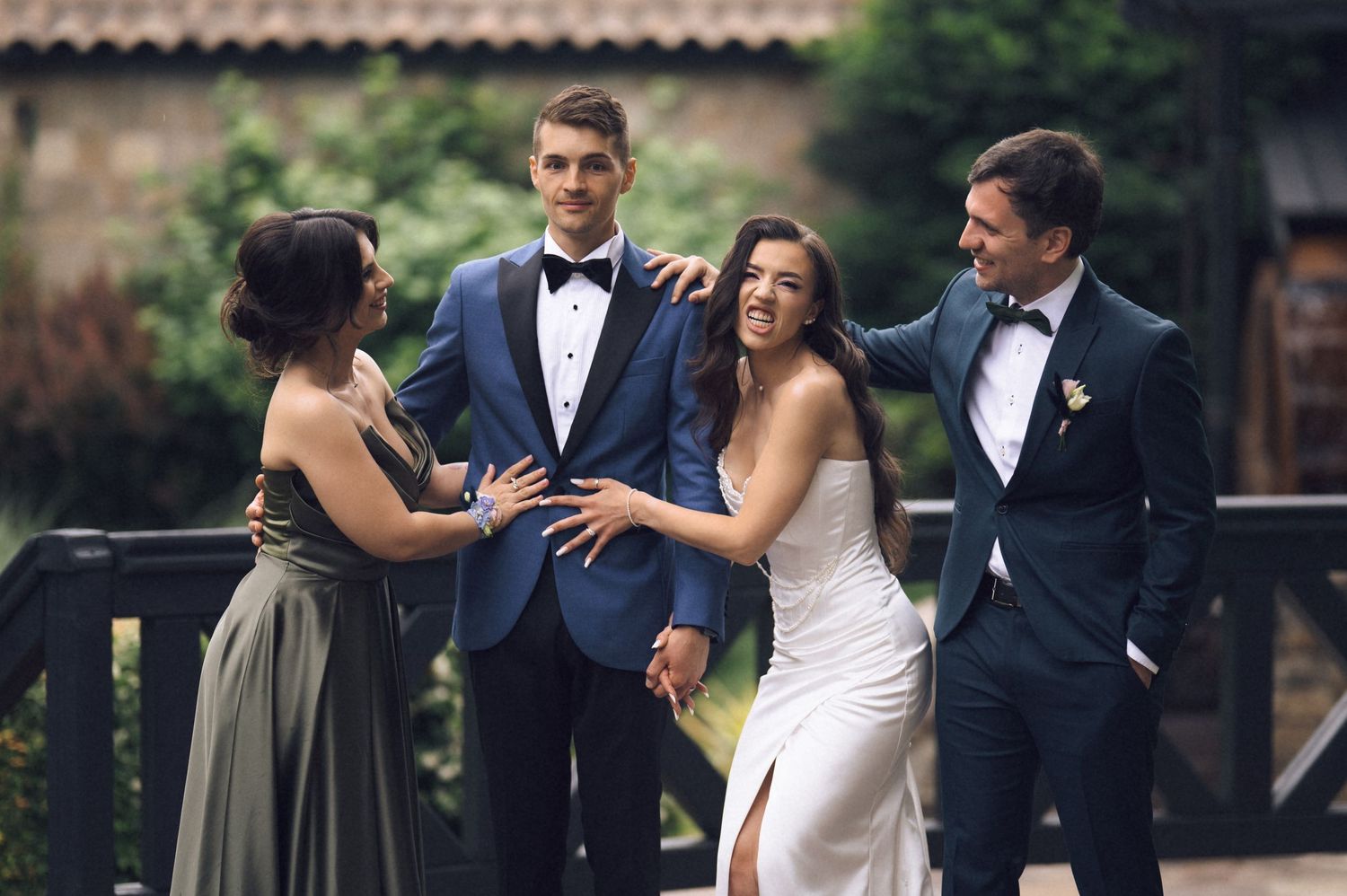 Wedding party of four posing outdoors, bride in white gown with groom in blue tuxedo, flanked by bridesmaid and groomsman.