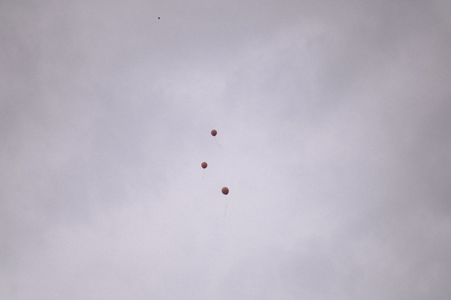 Three red balloons floating high in a cloudy gray sky, drifting upward on an overcast day.