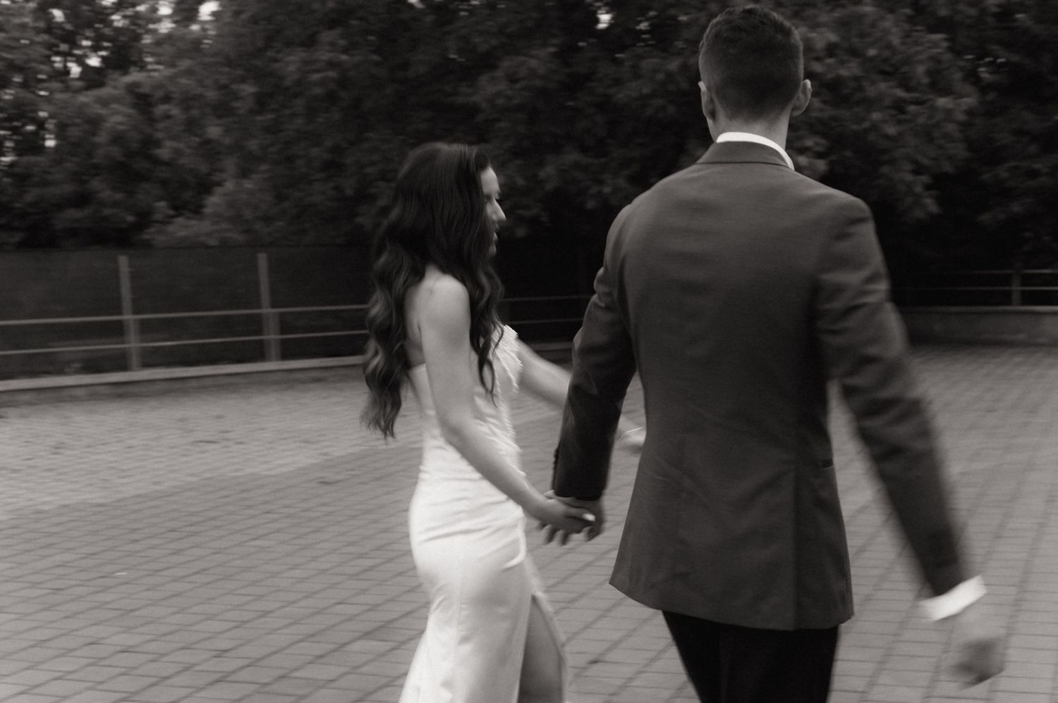 Bride in white fitted gown and groom in suit walk hand in hand outdoors in black and white wedding photo.