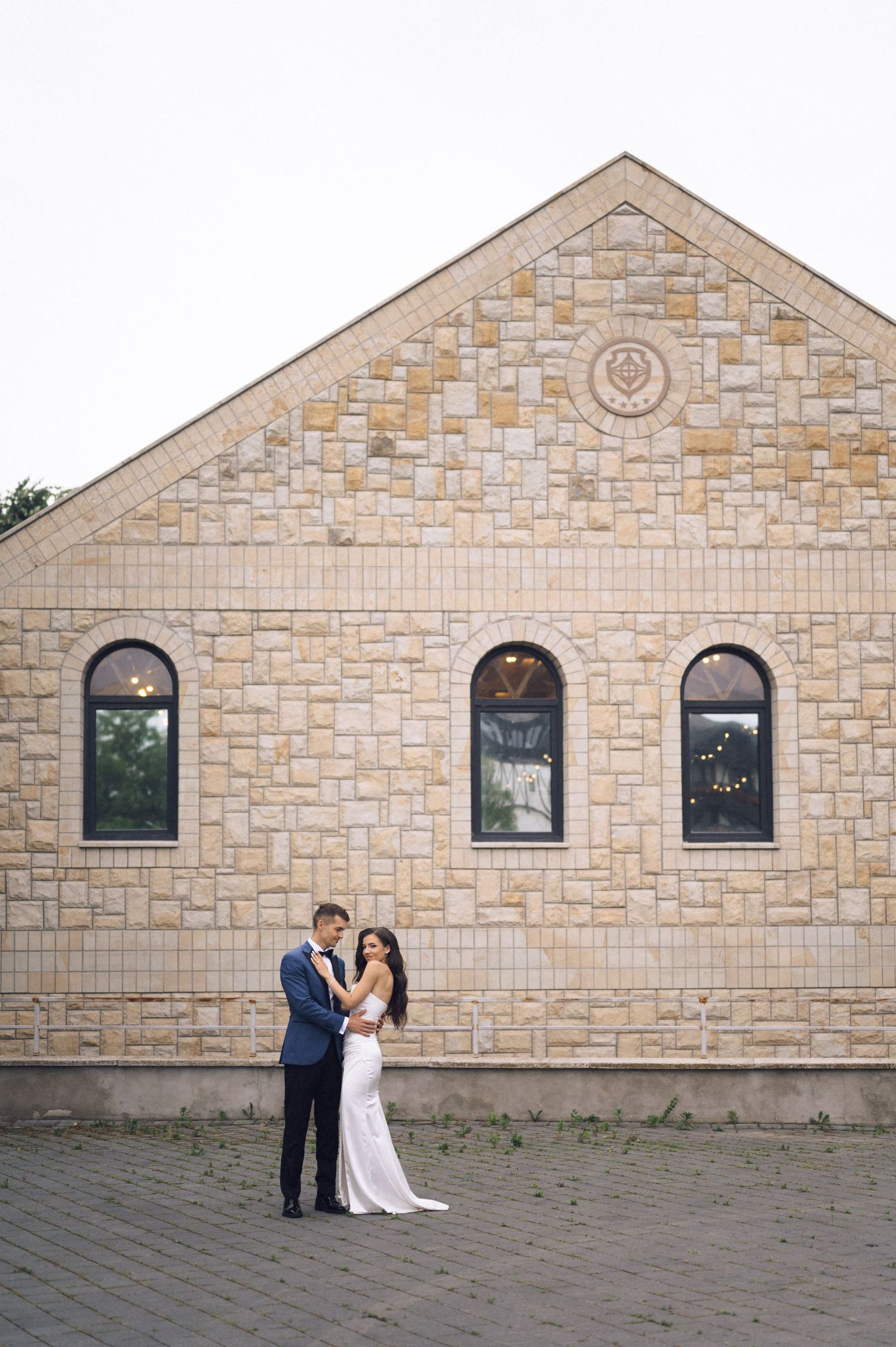 Bride and groom embracing in front of a rustic stone building with arched windows at their wedding venue.