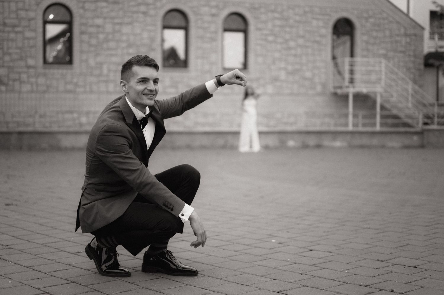 Black and white photo of a groom in a tuxedo crouching on a brick plaza, pointing toward a bride in the background.