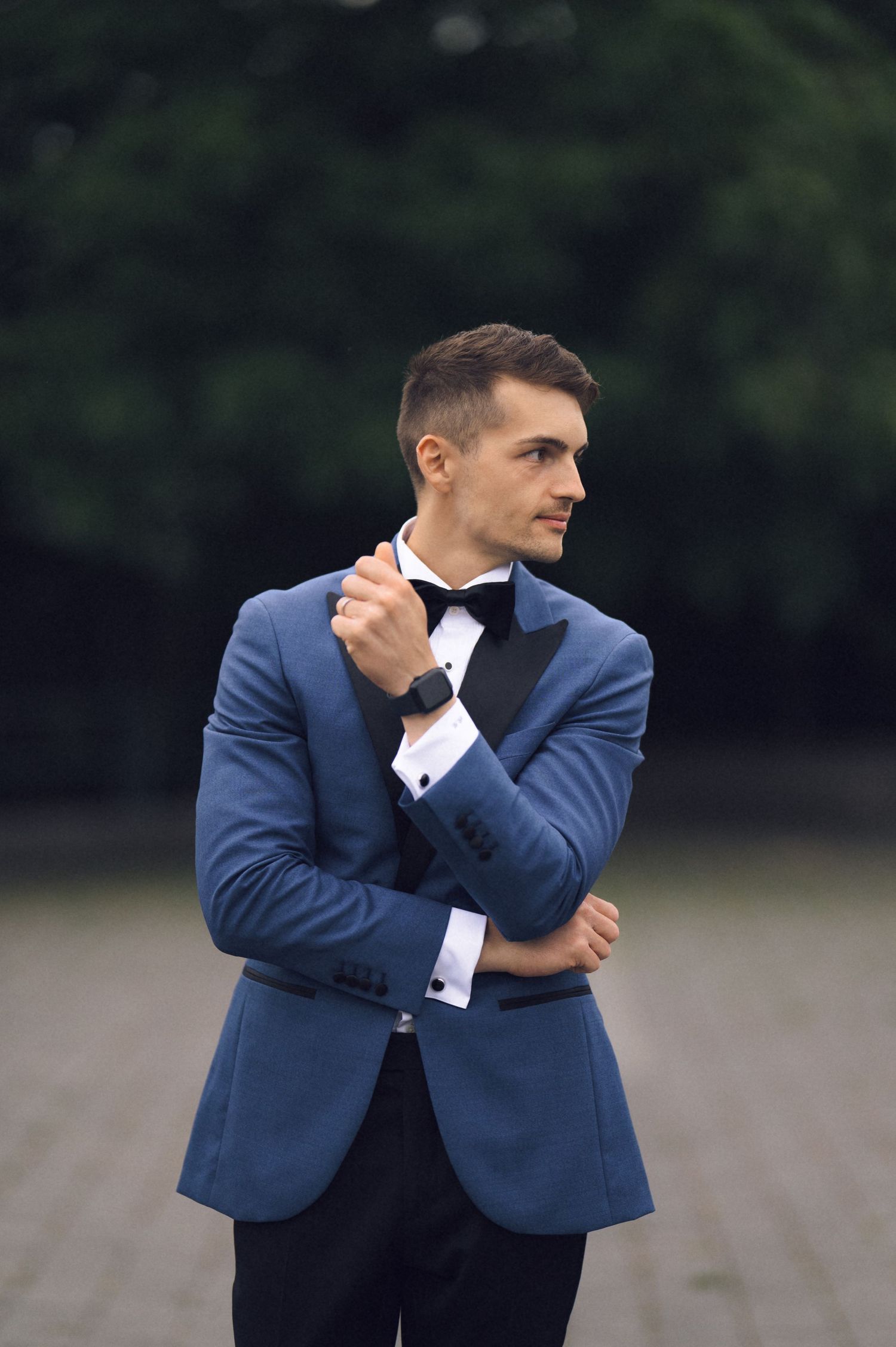 A stylish young man in a blue tuxedo jacket with black bow tie and smartwatch poses outdoors near green trees.