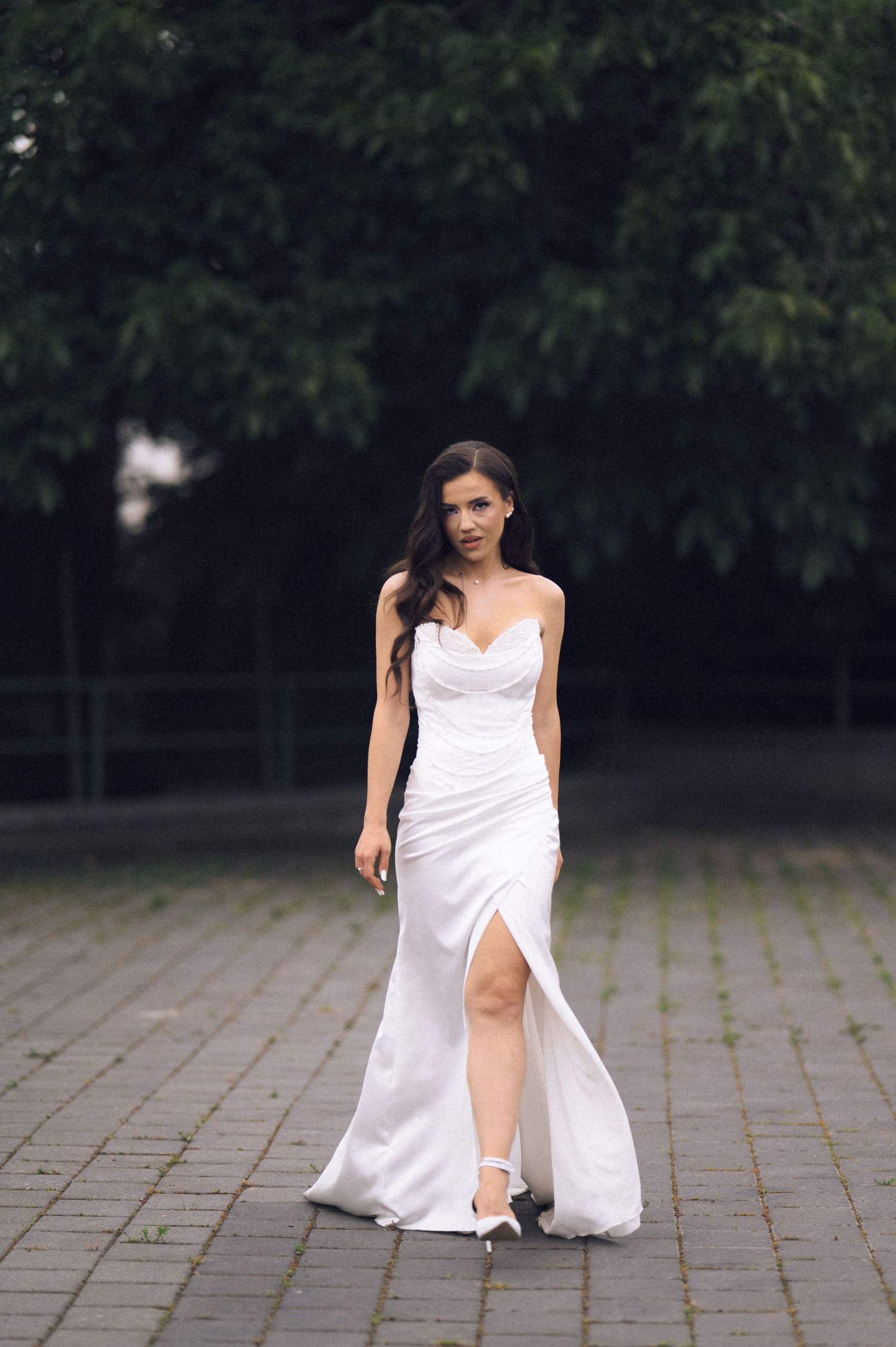 A bride in a white strapless slit gown walks gracefully on a paved path surrounded by lush green trees.