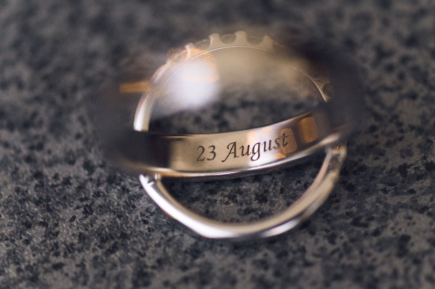 Two silver wedding rings engraved with the date 23 August, elegantly stacked on a gray stone surface.
