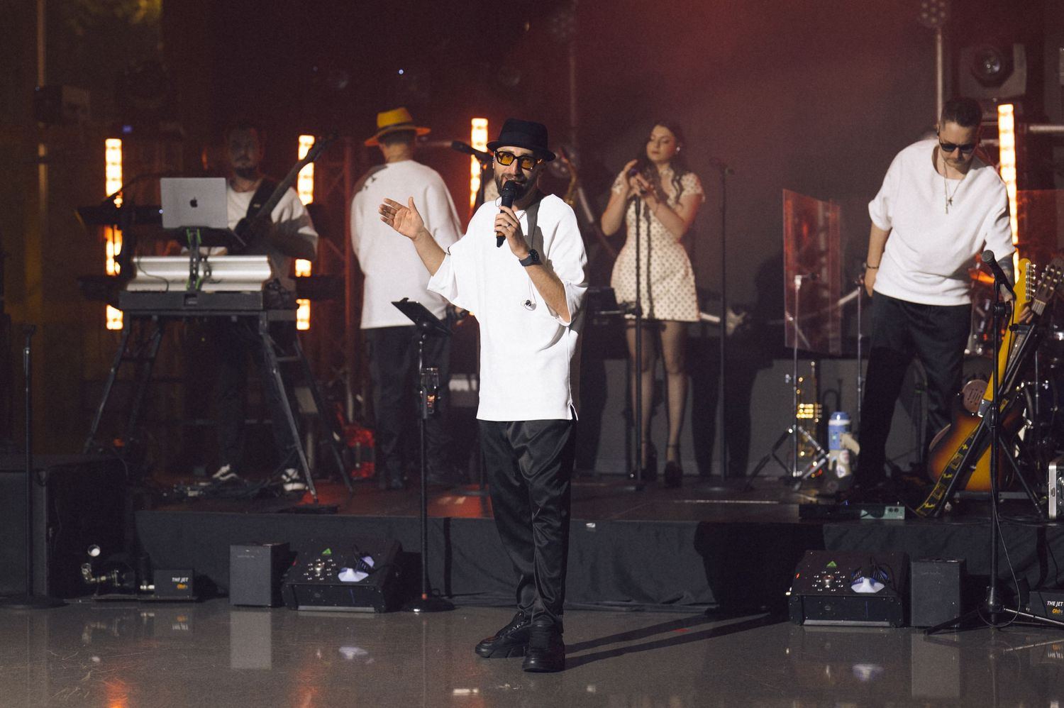 Live concert performance with singer holding microphone on stage, backed by band members and performers under warm lights.