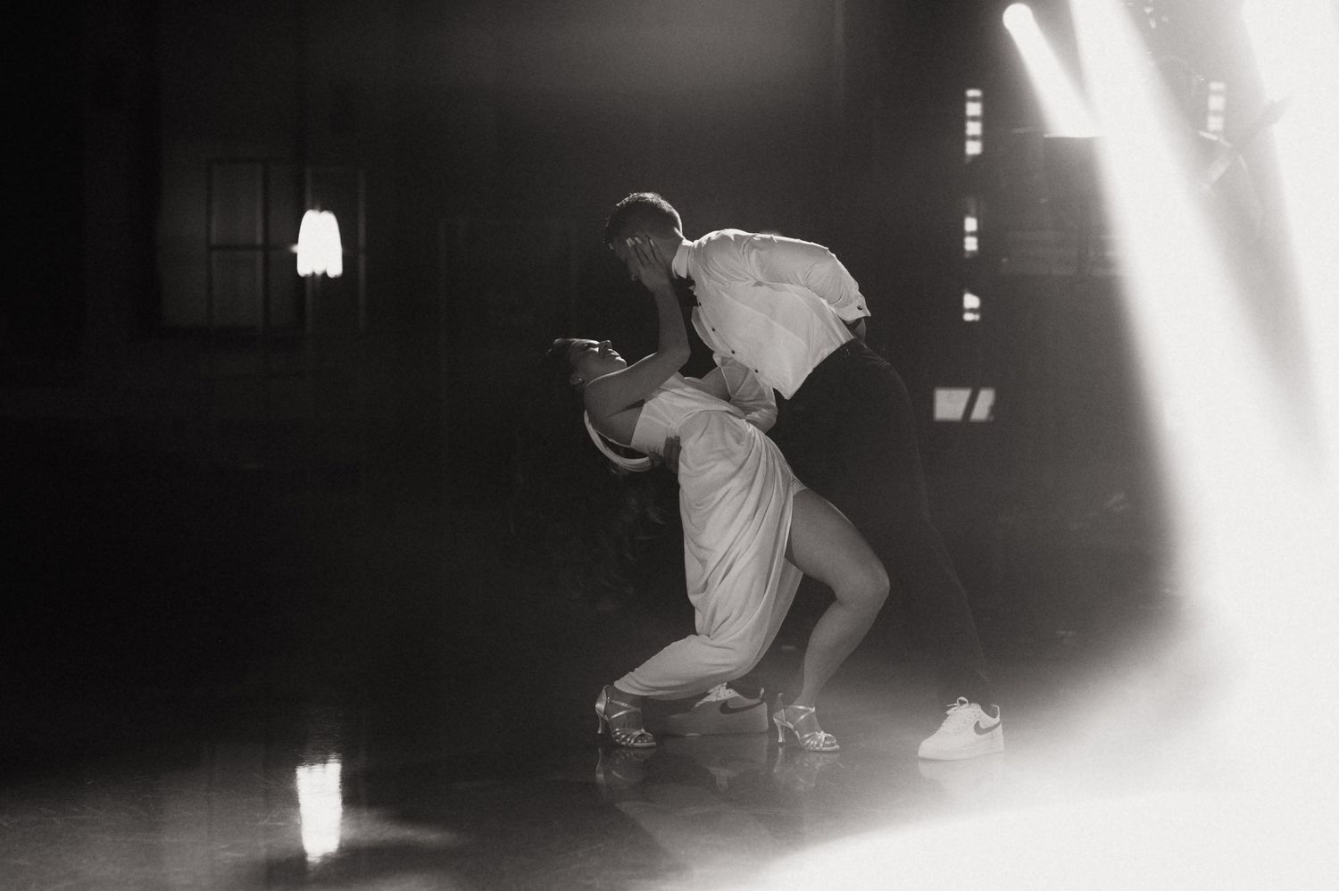 Two dancers perform an intimate dip in dramatic black and white lighting inside a dimly lit studio.
