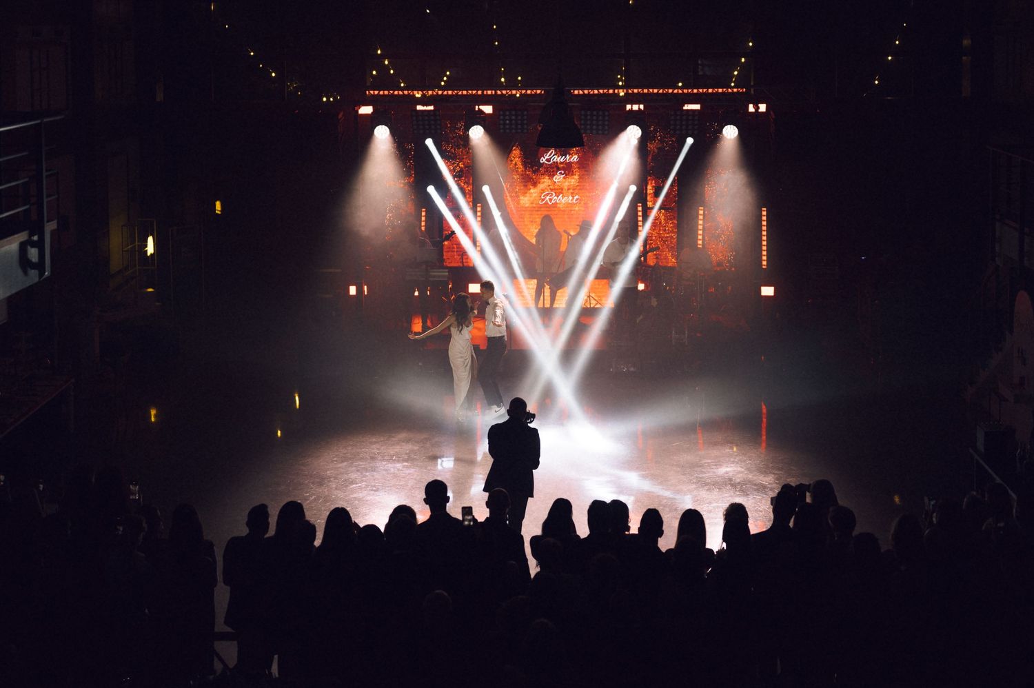 Silhouetted crowd watches a performer on a dramatic stage with bright spotlights and colorful backdrop at a live concert.