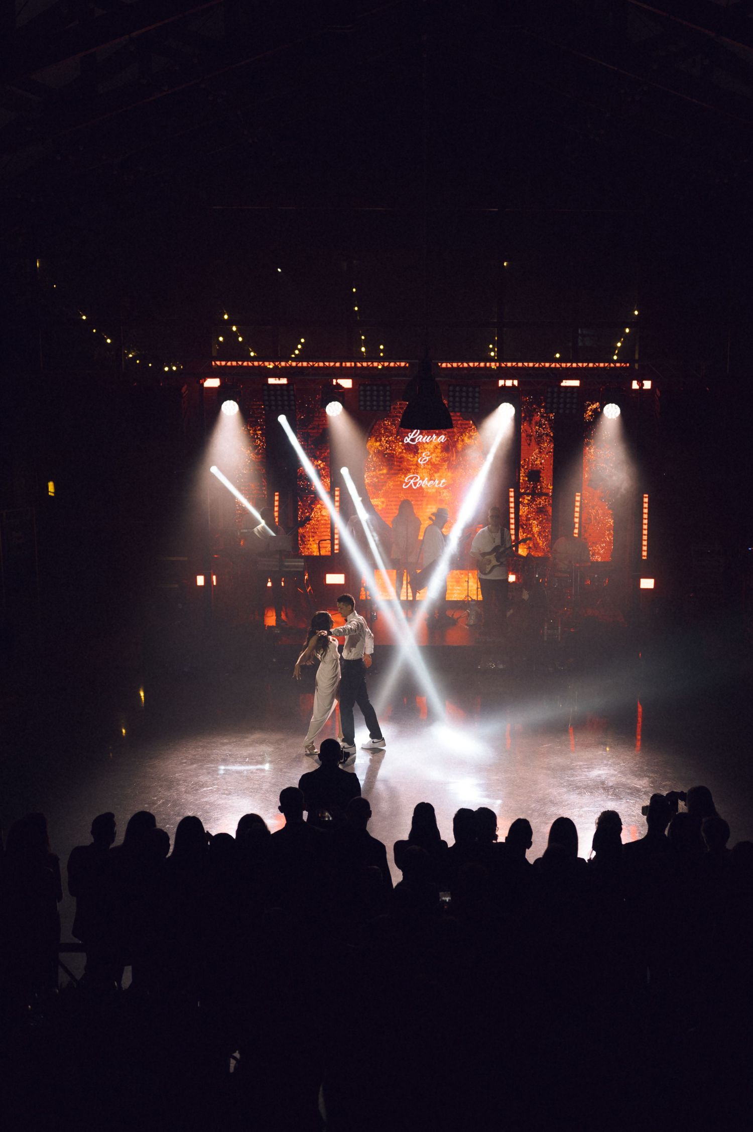 Two dancers perform on a dramatic stage with bright spotlights and warm orange backdrop before a silhouetted audience.