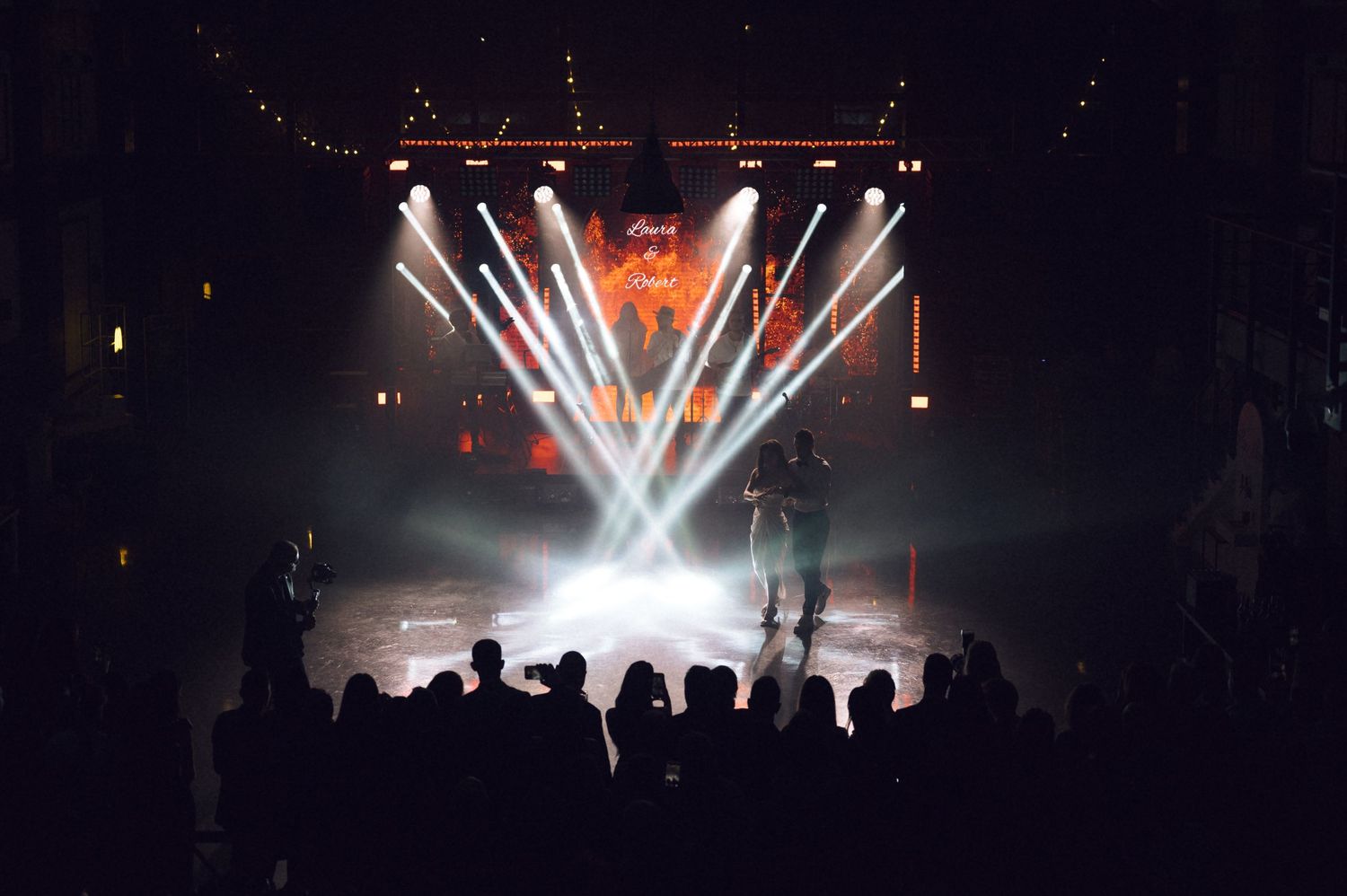 Silhouetted crowd watches a live concert with dramatic stage lighting and fog effects in a dark venue.