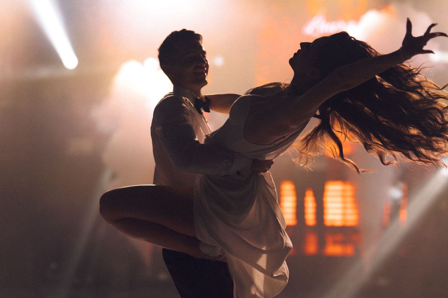Two dancers perform a dramatic lift on a smoky stage bathed in warm golden spotlight.