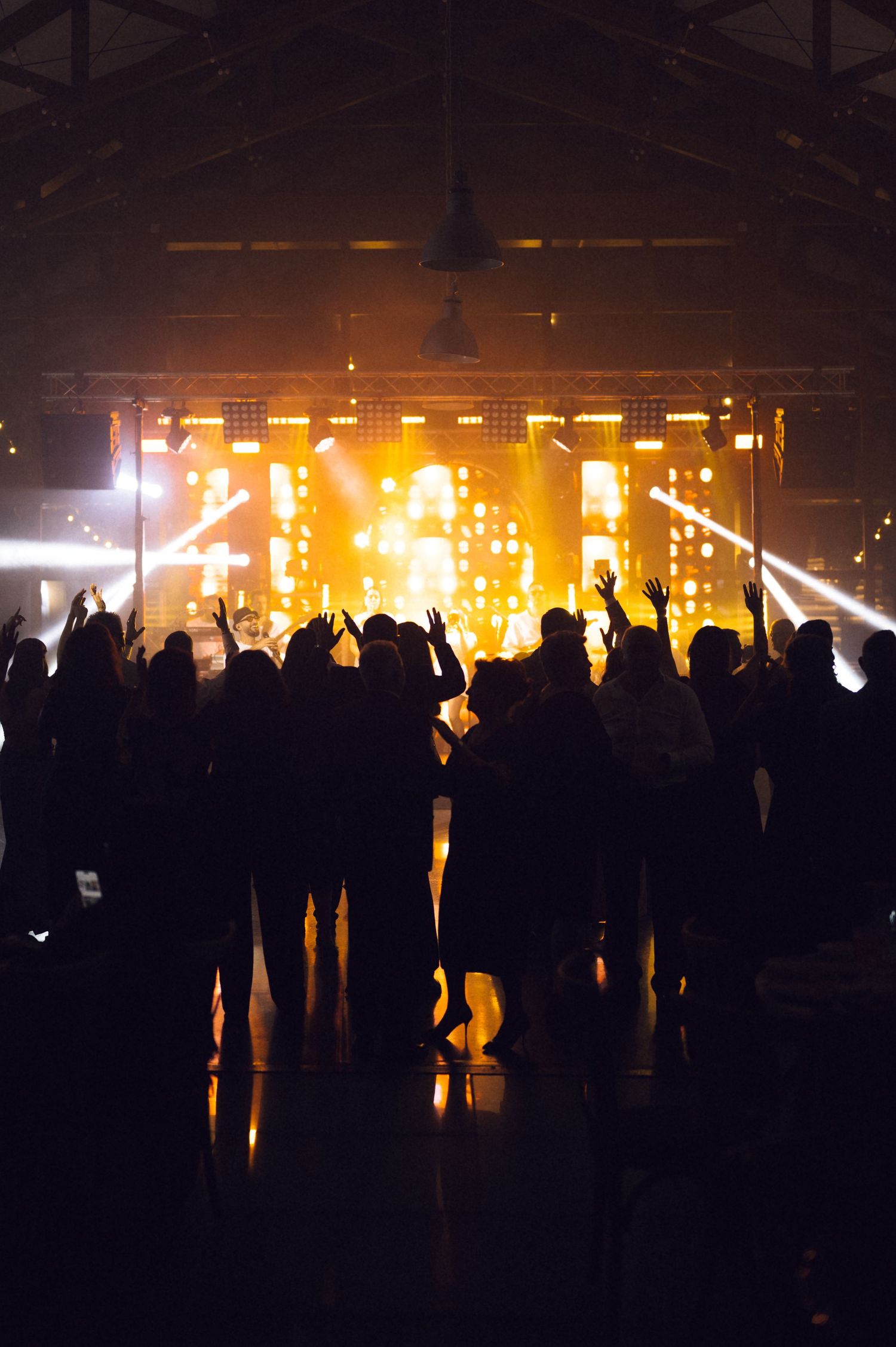 Silhouetted crowd dancing at a concert with vibrant yellow stage lights and LED screens in a dark venue.