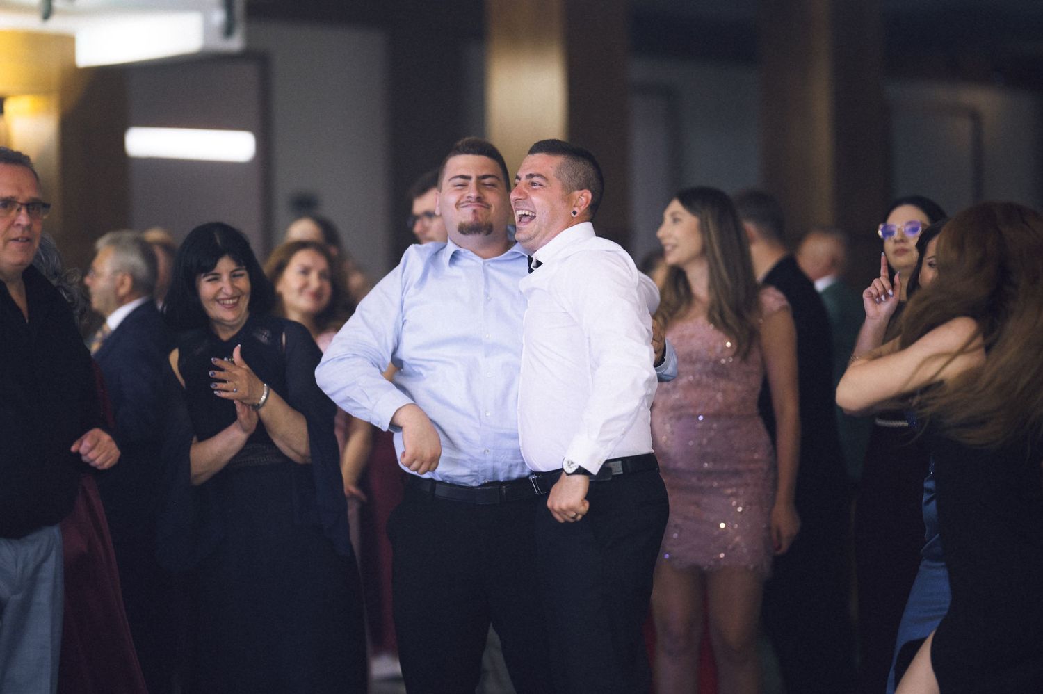 Two men dancing together at a formal event, surrounded by smiling guests in elegant attire.
