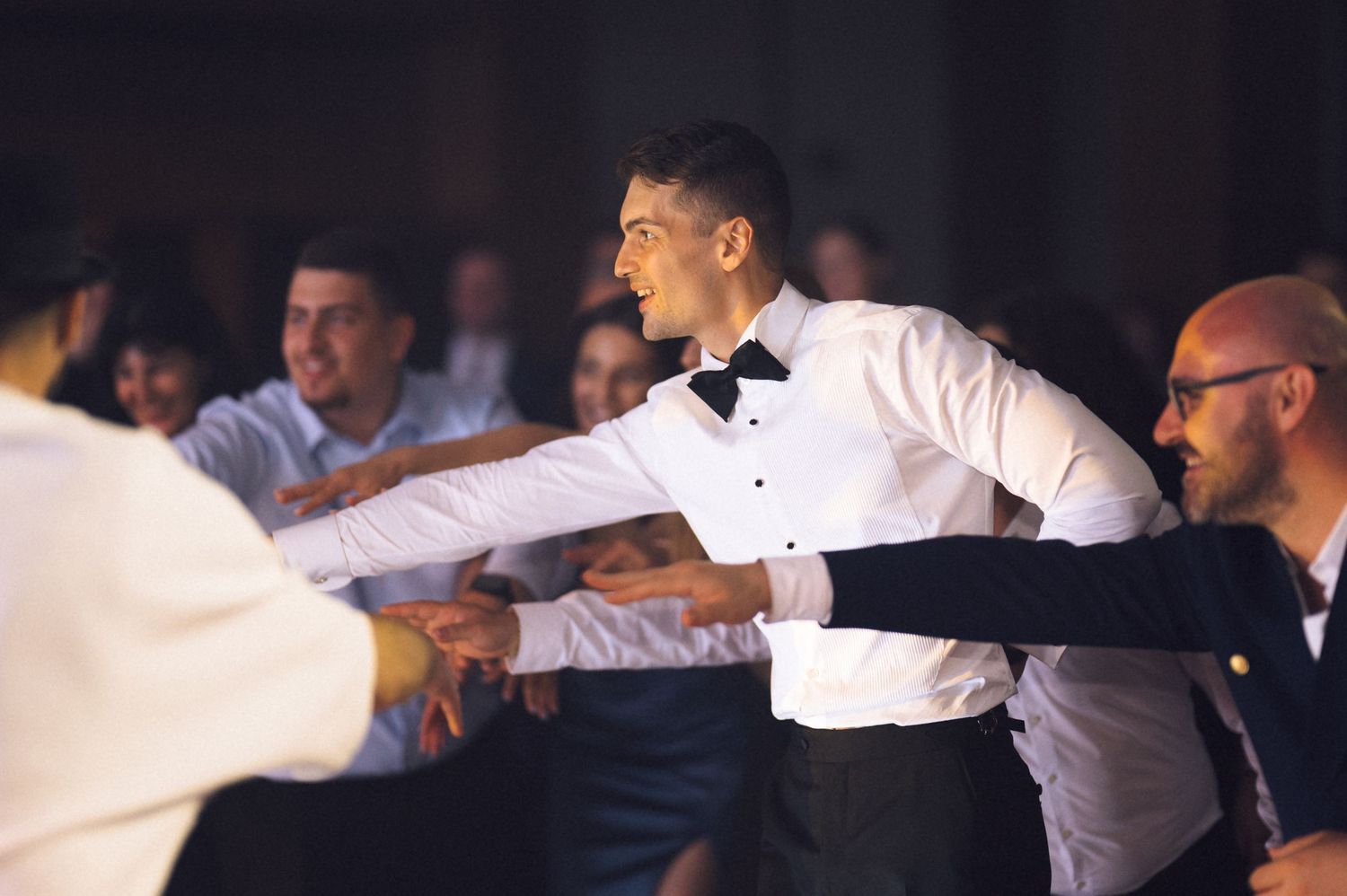 Man in tuxedo dancing joyfully at a formal event, reaching out to guests on a crowded dance floor.