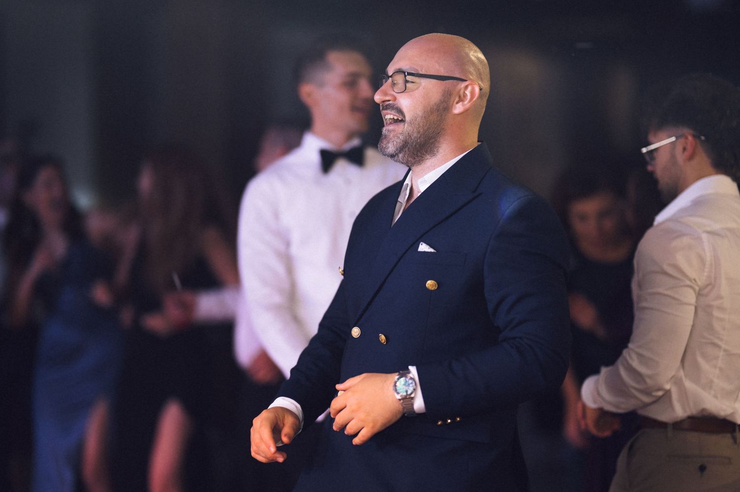 Bald man in navy double-breasted suit with gold buttons smiling at formal event.