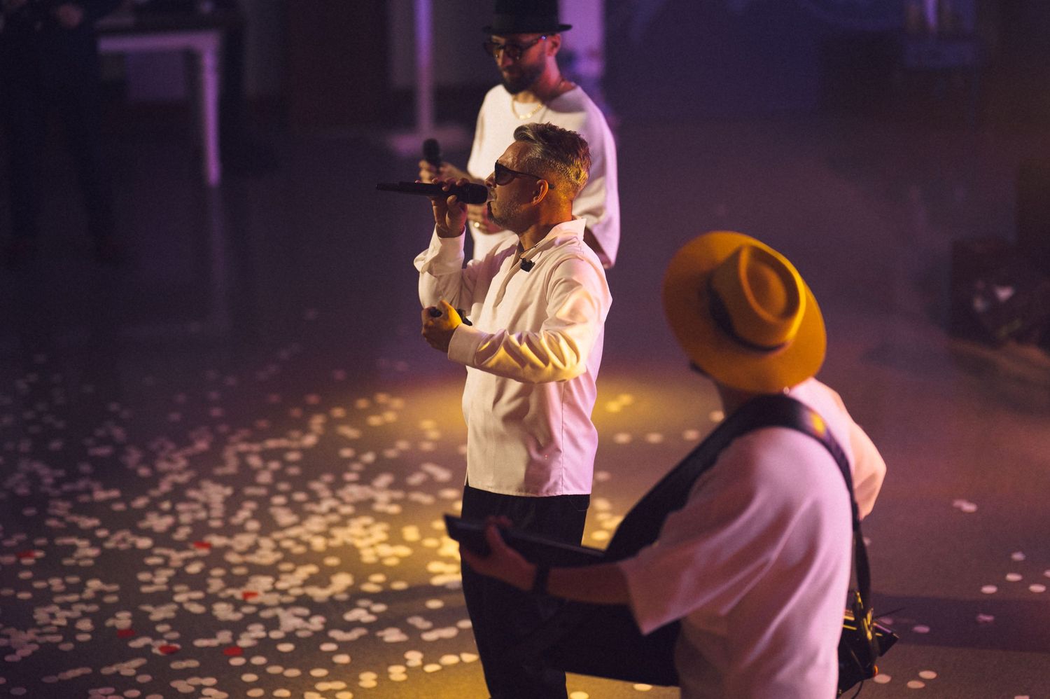 Three musicians performing on stage with confetti, one singing into a mic, another in a yellow hat playing guitar.