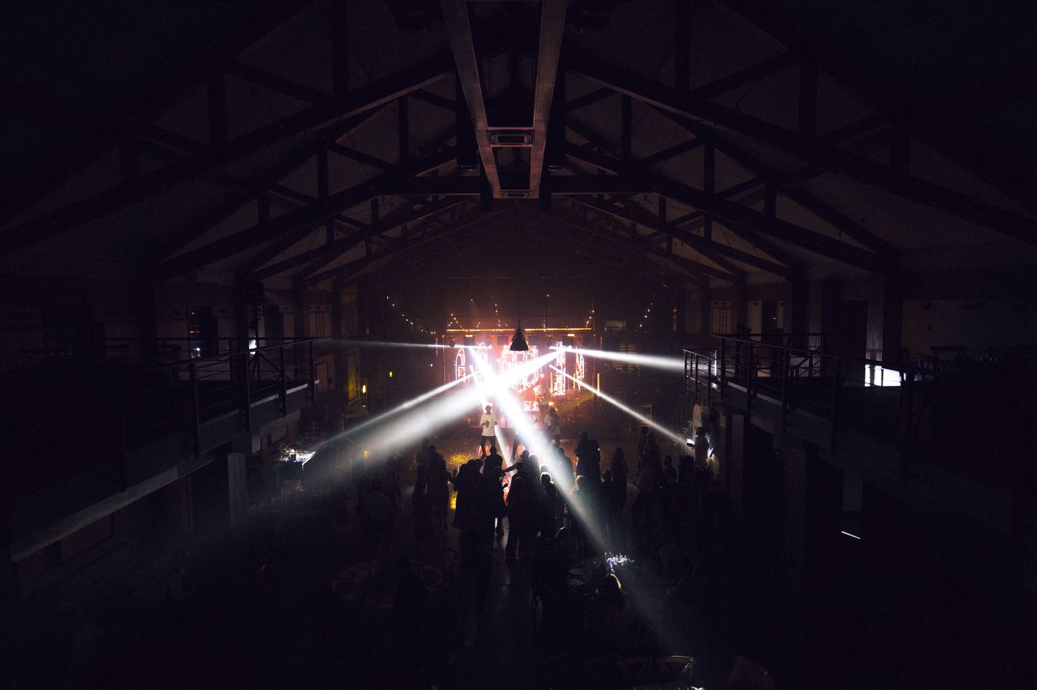 Dark warehouse venue with dramatic light beams illuminating a crowd during a live music or rave event.