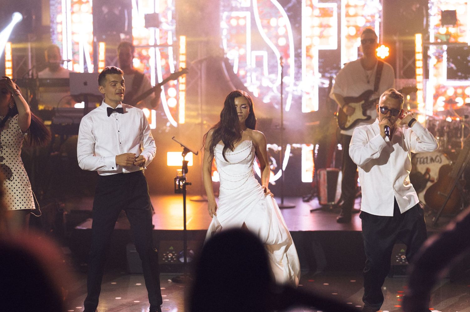 Three performers in elegant white outfits singing on a glamorous concert stage with bright lights.