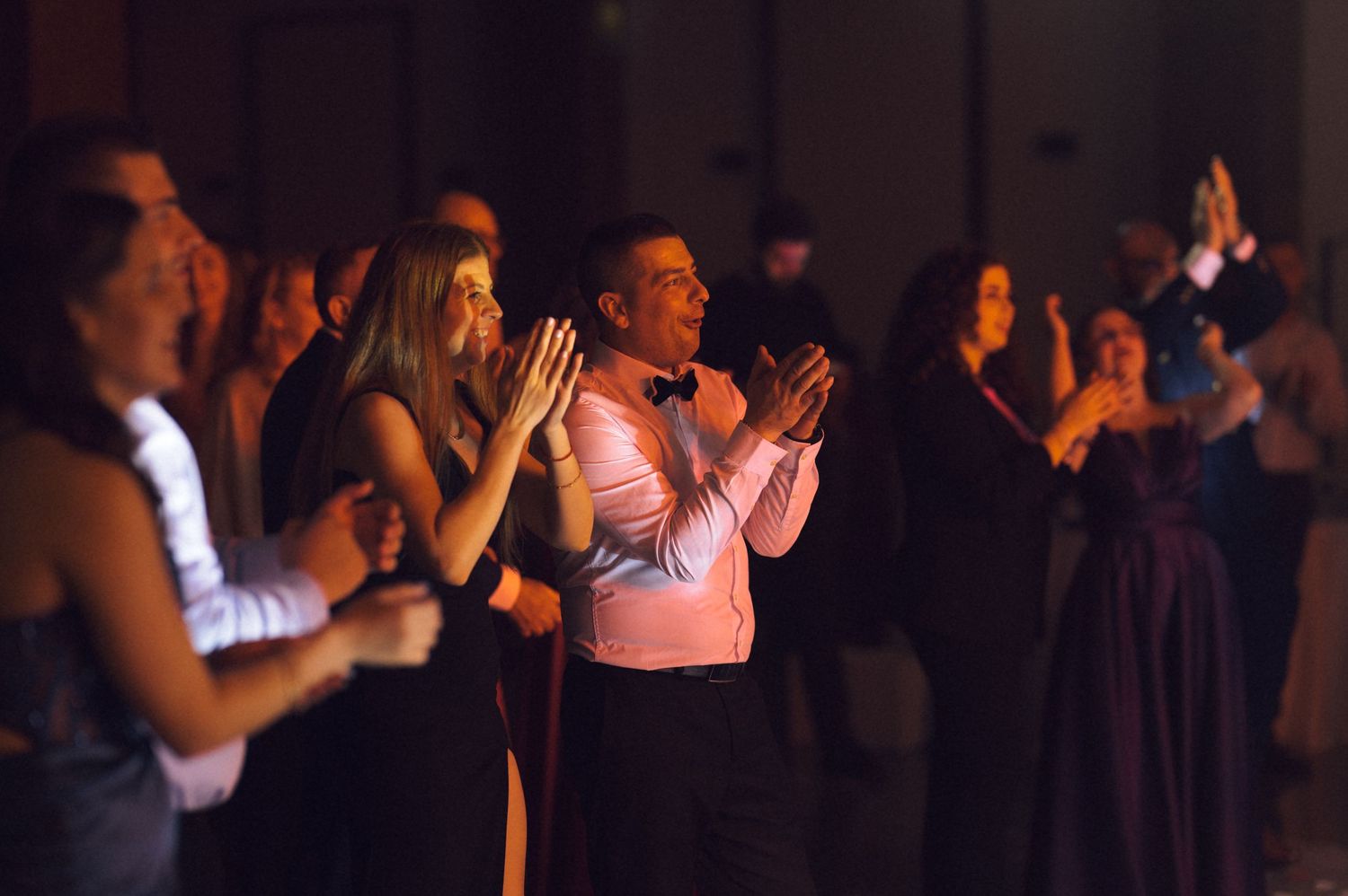 Elegantly dressed guests clapping and celebrating at a formal gala event in a dimly lit ballroom.
