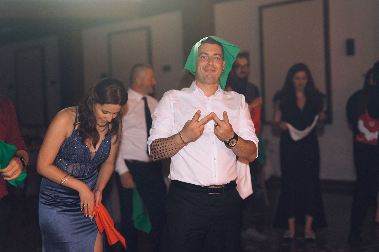Man dancing with green napkin on head at wedding reception party with guests in background.