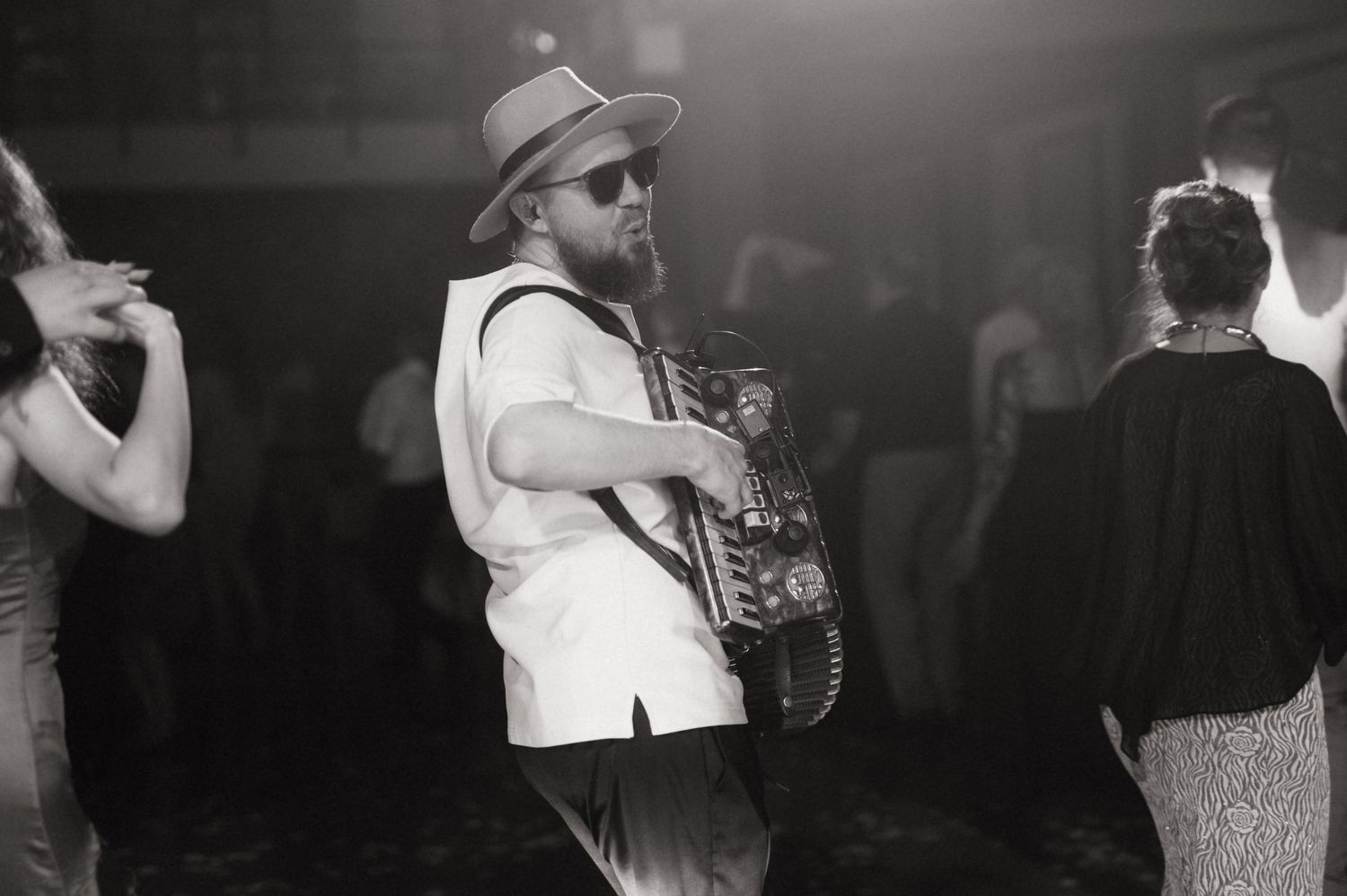 Black and white photo of a saxophonist in a wide-brim hat performing at a lively dance event.