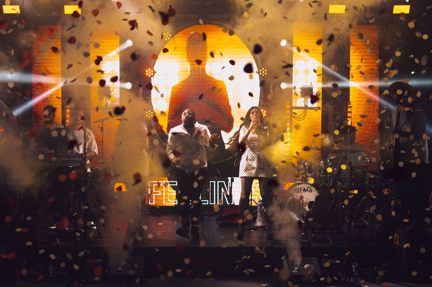 Performers on stage with golden confetti falling during an energetic live concert under warm stage lighting.