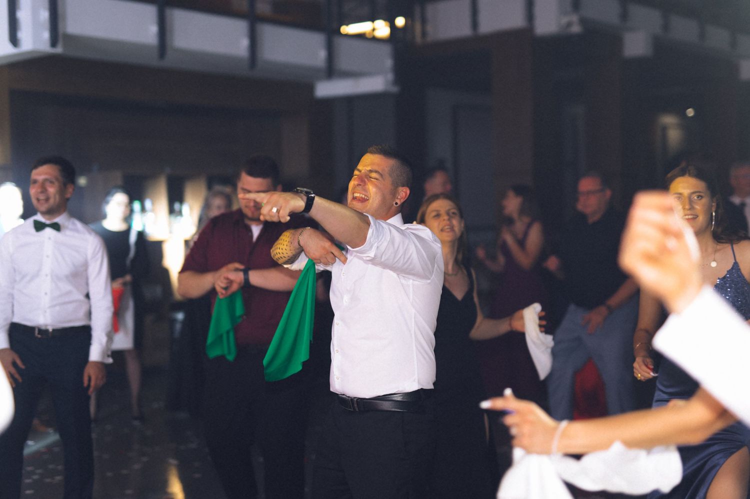 Guests dancing energetically at a wedding reception in a dimly lit venue, with a man in white shirt leading the fun.