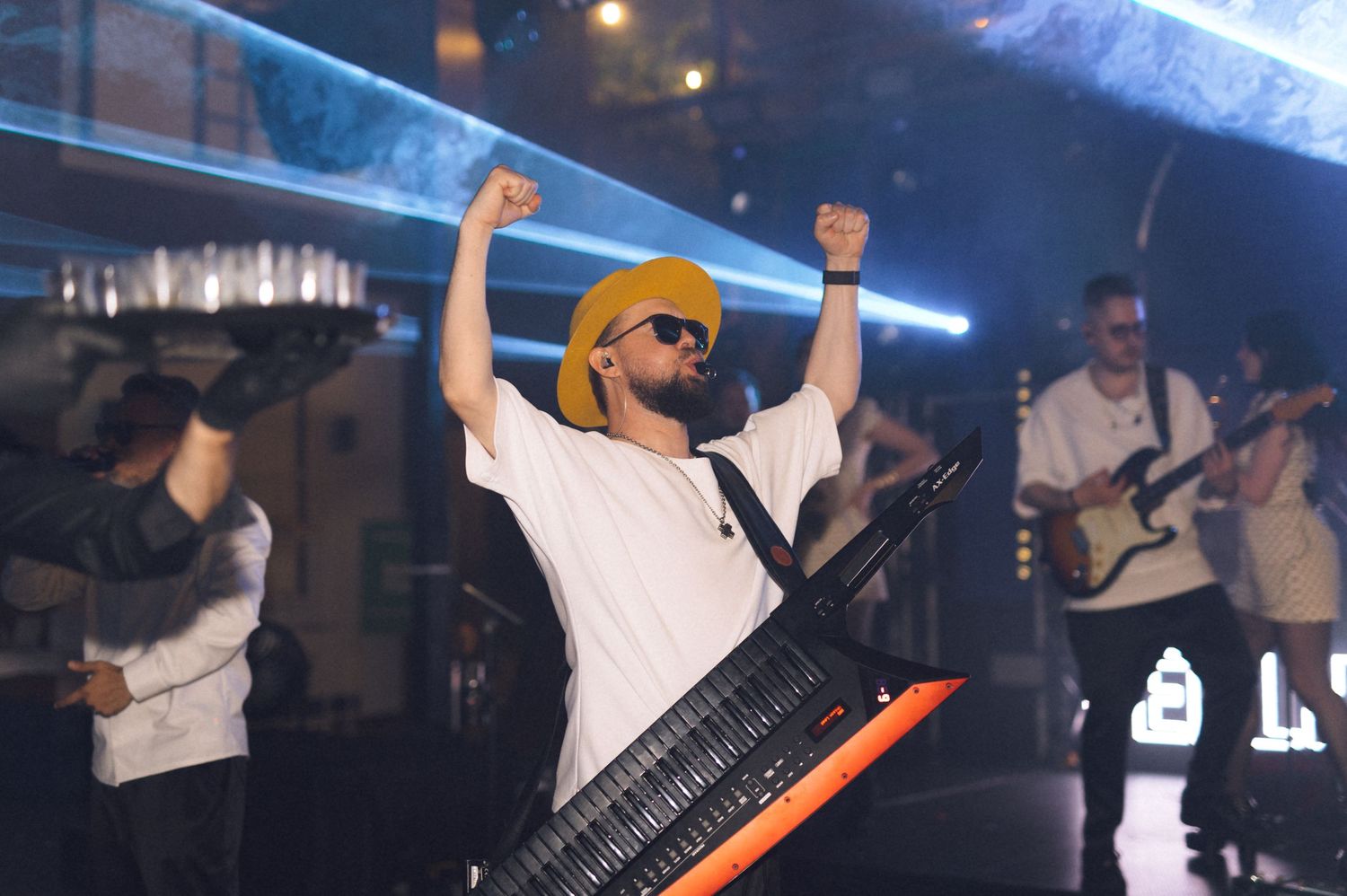 Excited musician in yellow hat and sunglasses raises fists while playing keyboard on stage with band.