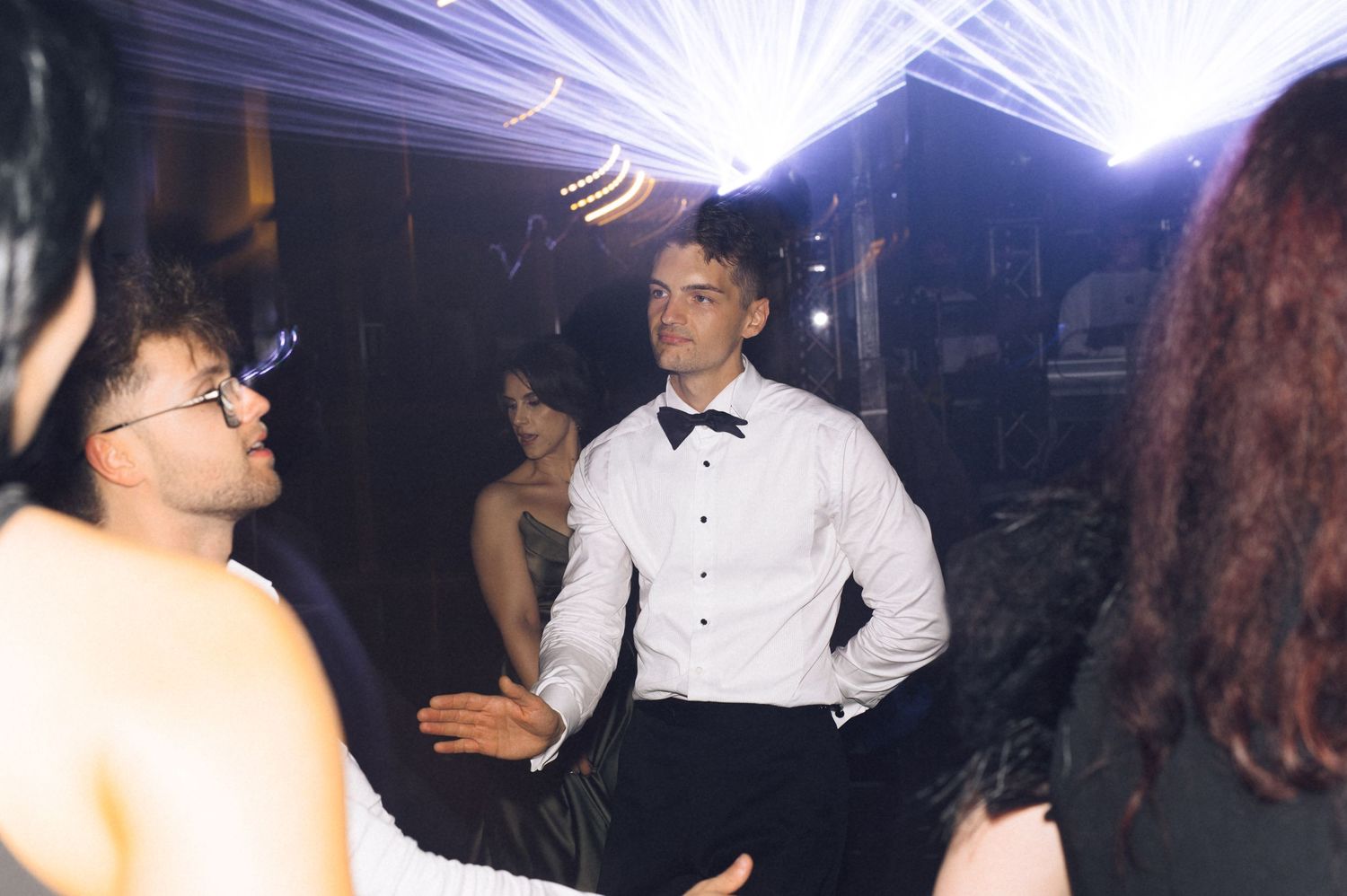 Young man in white dress shirt and bow tie dancing at a formal event with guests in elegant attire.