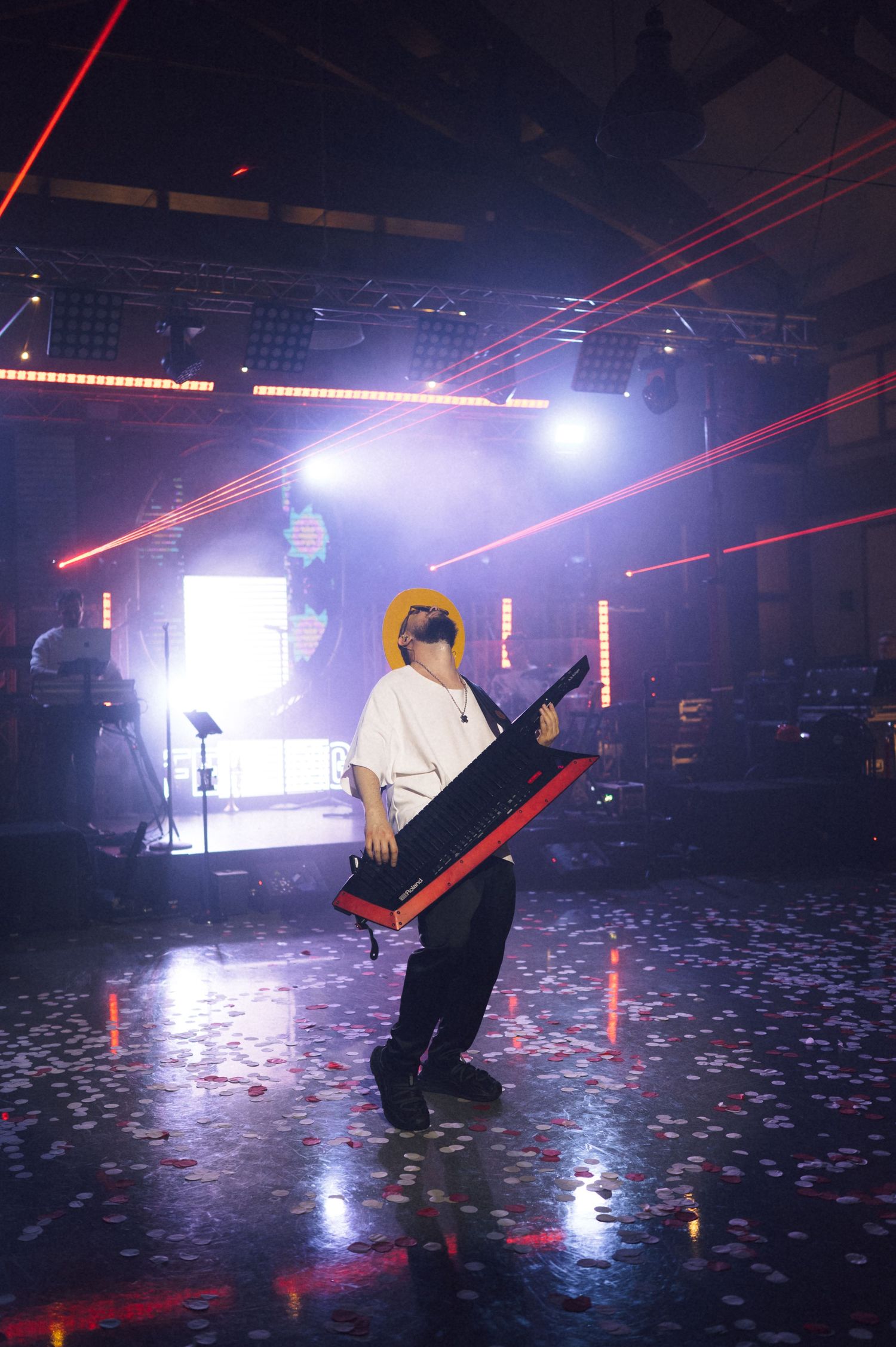 Musician plays a keytar on stage surrounded by red laser lights and confetti at a live concert event.