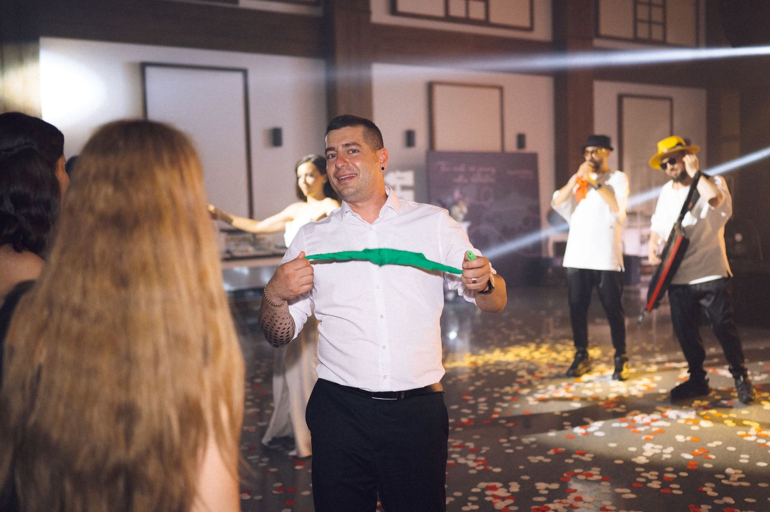 Man in white shirt stretches green resistance band at a festive indoor celebration with confetti and guests.