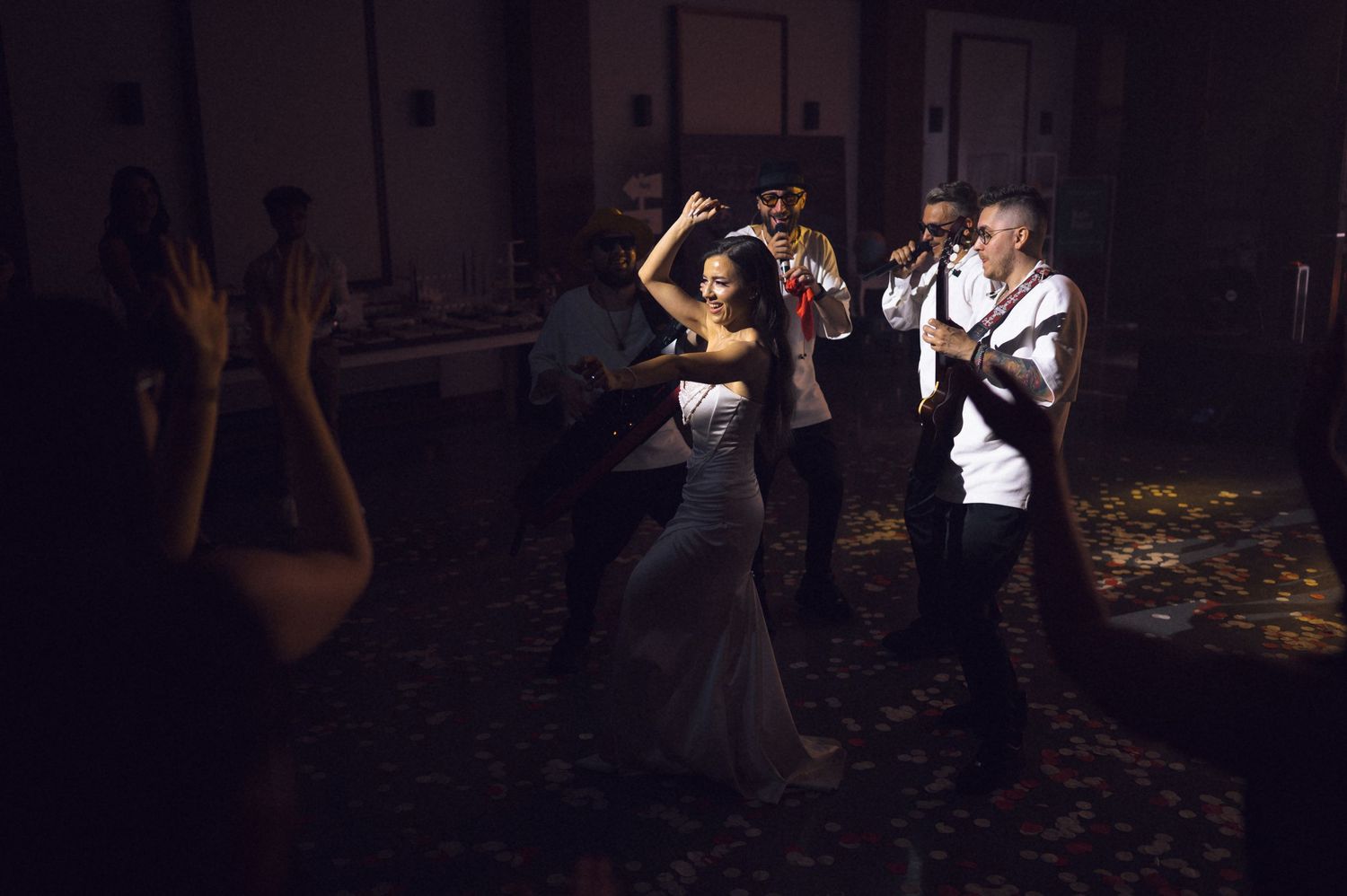 Bride in white gown dancing with guests as a live band performs at a dimly lit wedding reception.