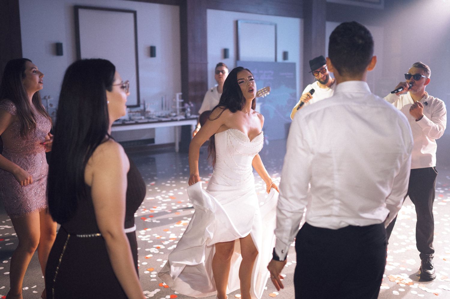 Bride in white wedding dress dancing joyfully with guests at elegant wedding reception with confetti.