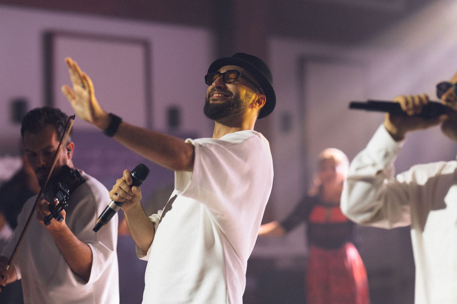 Man in white outfit singing on stage with microphone, arm raised, performing live with backup singers behind him.