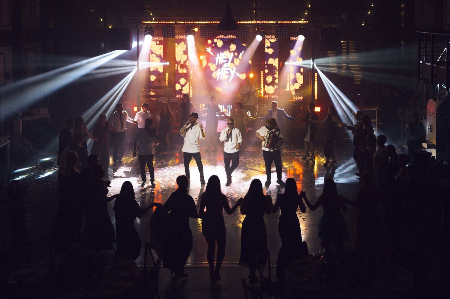 Silhouetted crowd watches performers on a brightly lit concert stage with dramatic spotlights and warm golden lighting.