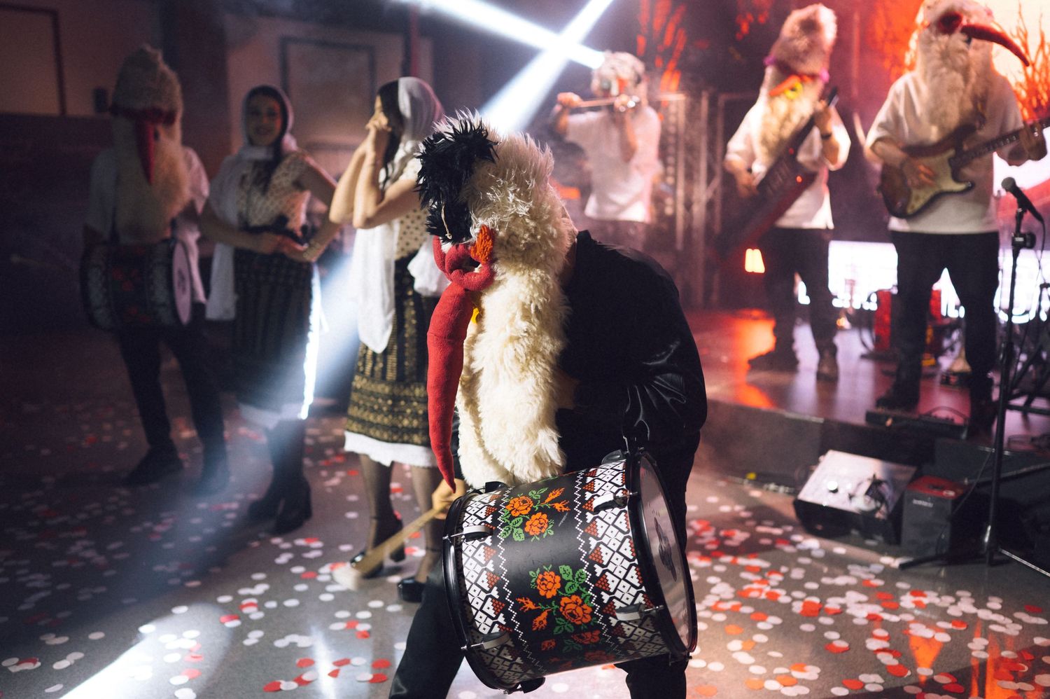 Performer in traditional folk costume with drum on stage during vibrant live music event with band and colorful lighting.