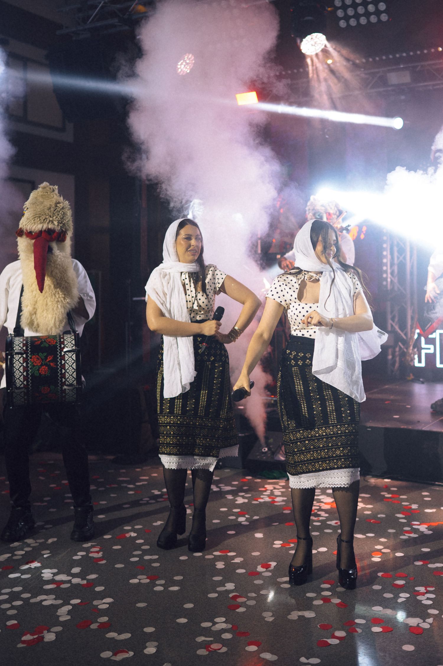 Women in traditional Romanian folk costumes perform on a smoke-filled stage with colorful confetti and bright lights.