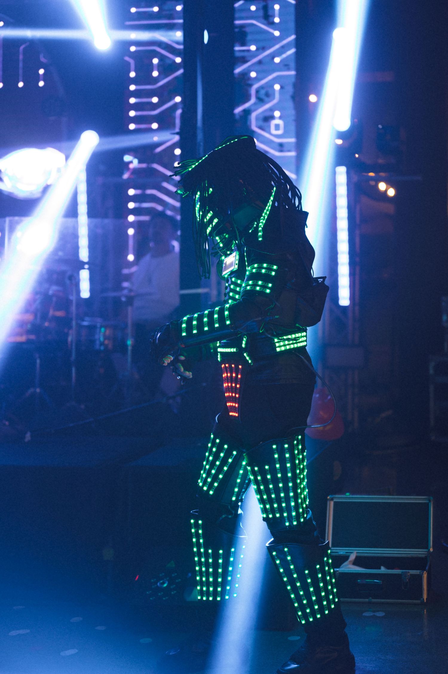 Performer in glowing green LED suit on stage with neon lights and circuit board backdrop at a futuristic event.