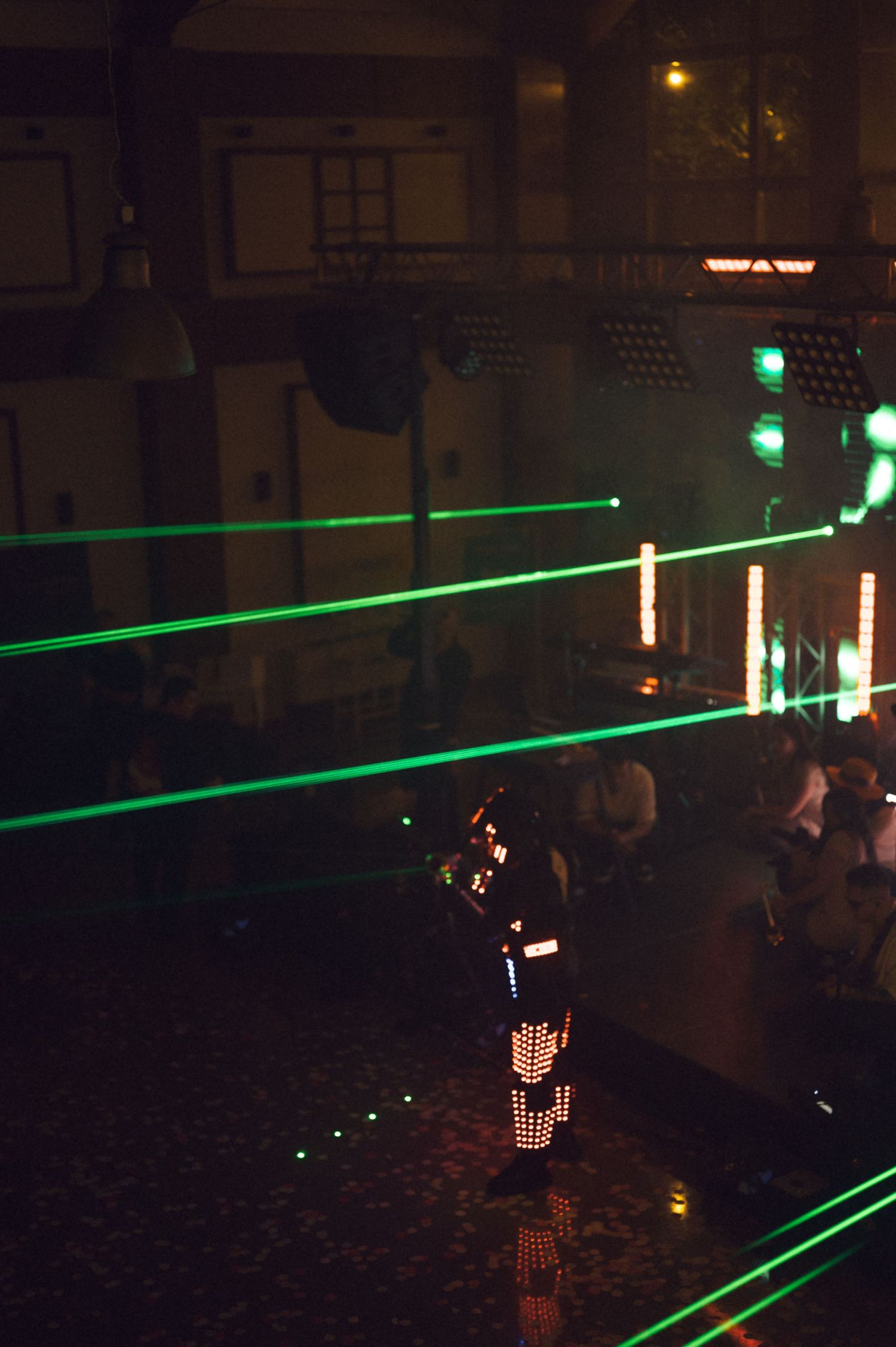 Performer in LED suit with green laser beams on dark nightclub stage during live show.