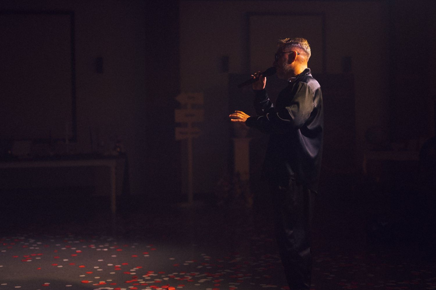 A person stands in a dimly lit room with dramatic lighting and red confetti scattered on the floor.