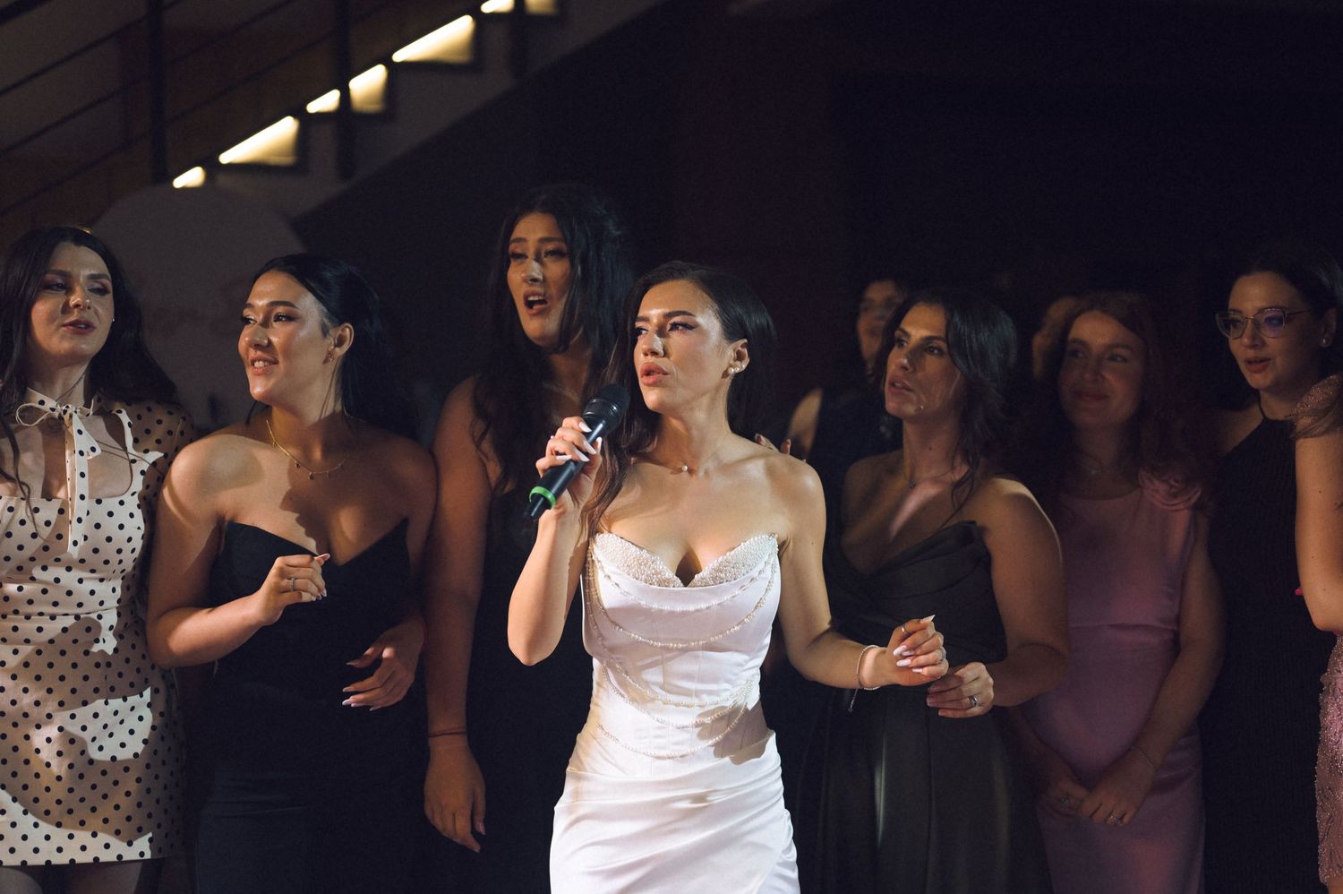 A woman in a white dress sings into a microphone surrounded by elegantly dressed women at a formal event.