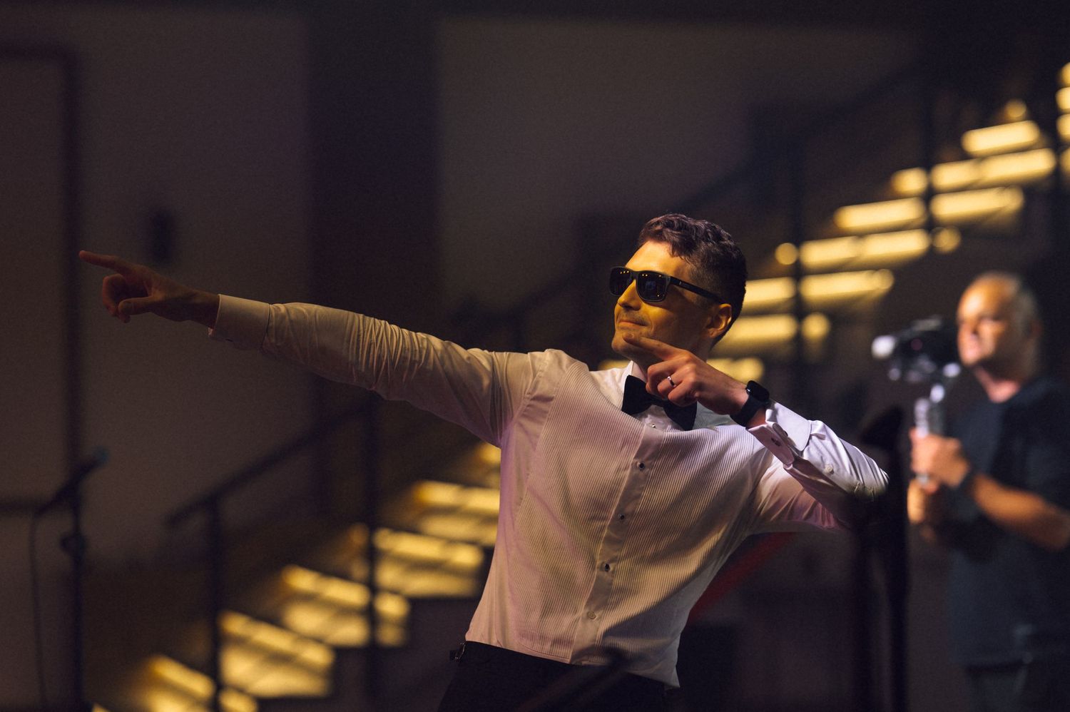 Man in white shirt and bow tie with sunglasses pointing dramatically on stage during a performance.