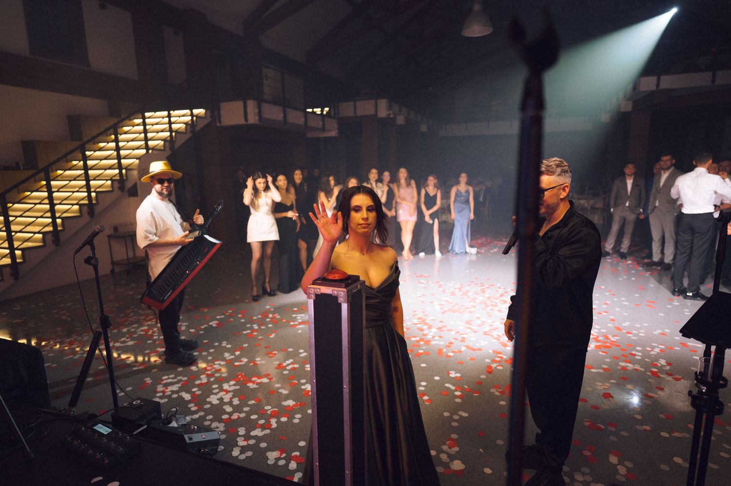 A woman in a black gown waves on a rose petal-covered stage with performers and staircases in background.
