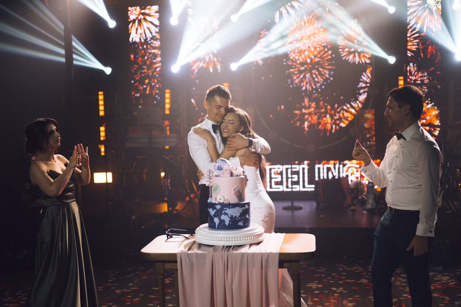 Couple embracing behind wedding cake at elegant reception with fireworks display and neon sign backdrop.