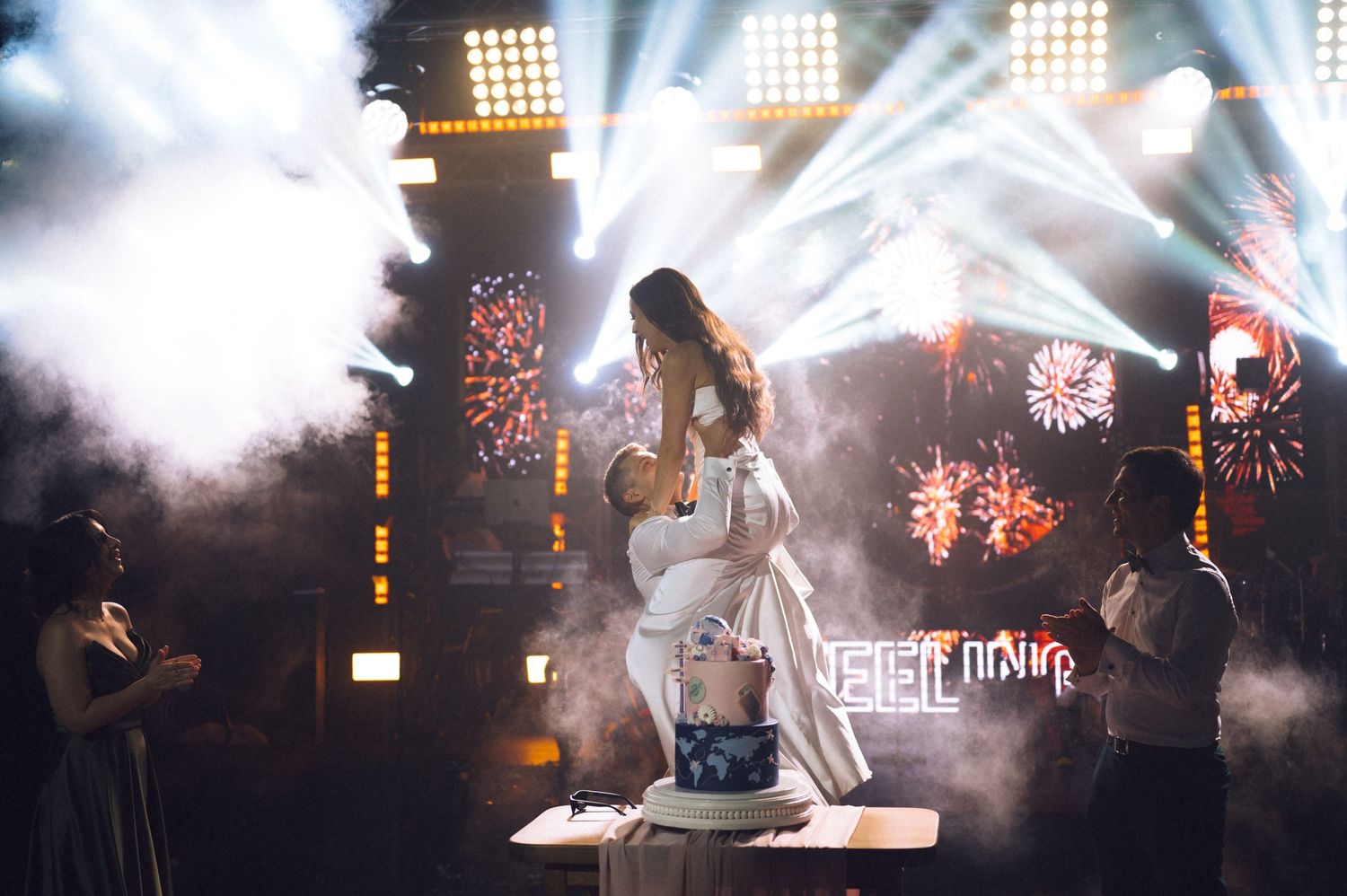 A performer in white dress stands on stage amid smoke, fireworks, and dramatic lighting at a live event.