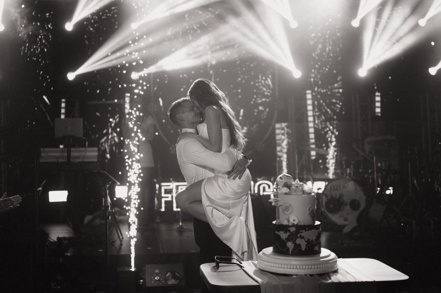Black and white wedding photo of couple embracing on stage with sparklers and dramatic lighting, cake in foreground.