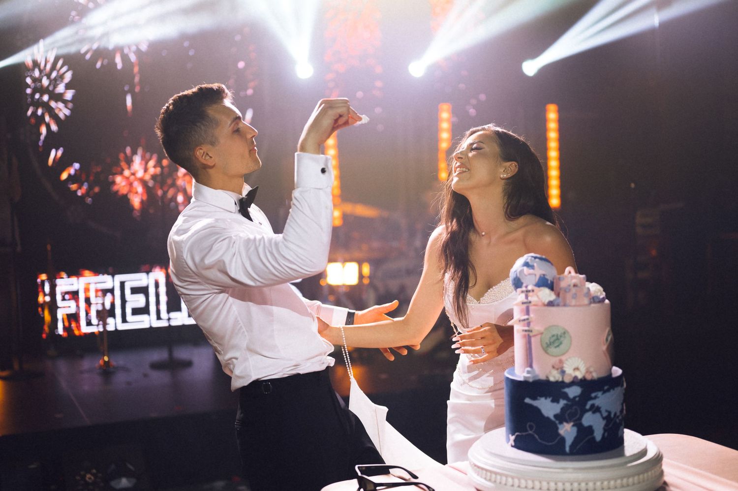 Bride and groom cutting their wedding cake on a decorated stage with fireworks and stage lights in the background.