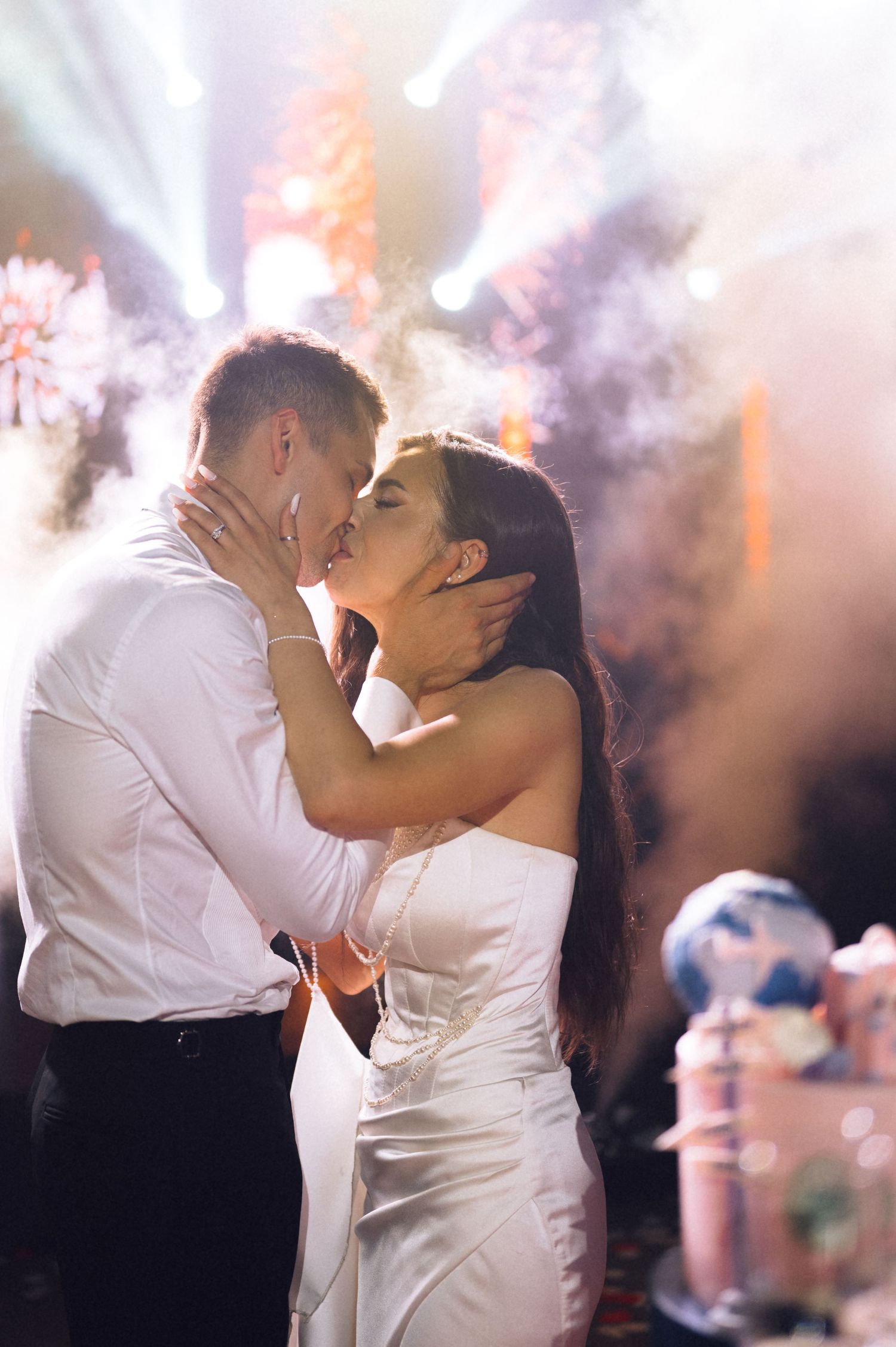 A couple shares a romantic kiss in wedding attire, surrounded by dramatic lighting and smoke effects near a decorated cake.