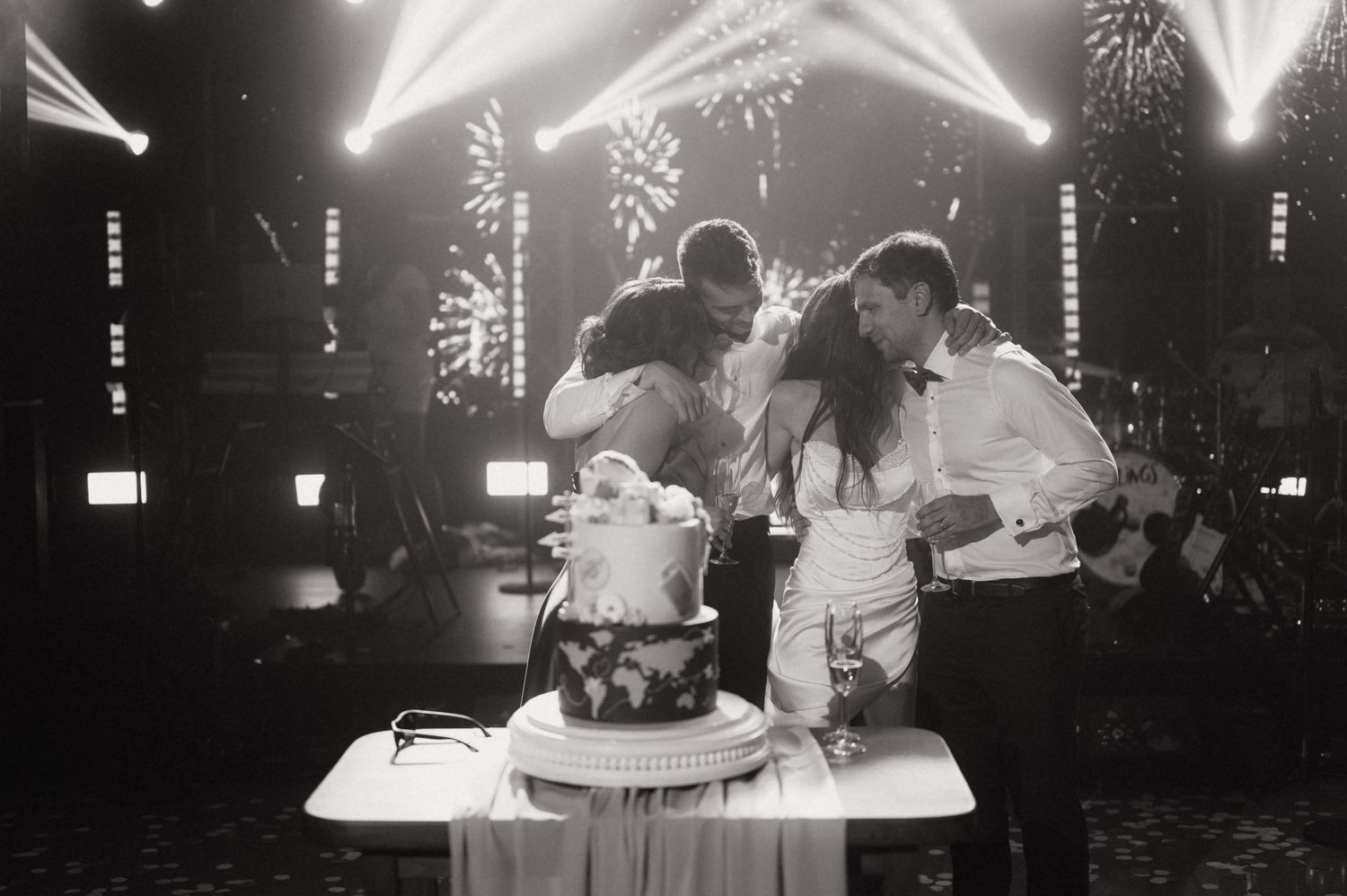 Black and white wedding photo of couple kissing over tiered cake with fireworks and stage lights in background.