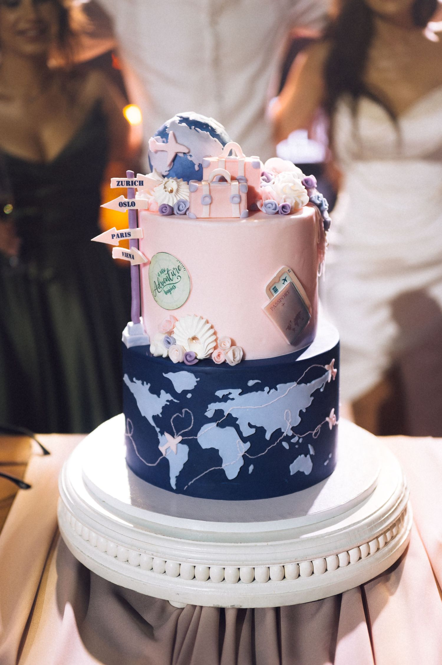 Two-tier travel-themed wedding cake with world map base, pink top tier, luggage toppers, and city destination signs.