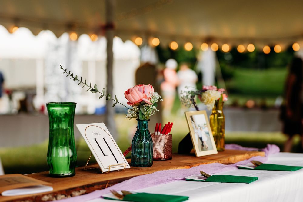 Elegant wedding reception table with green vases, pink roses, table numbers, and string lights under a white tent.